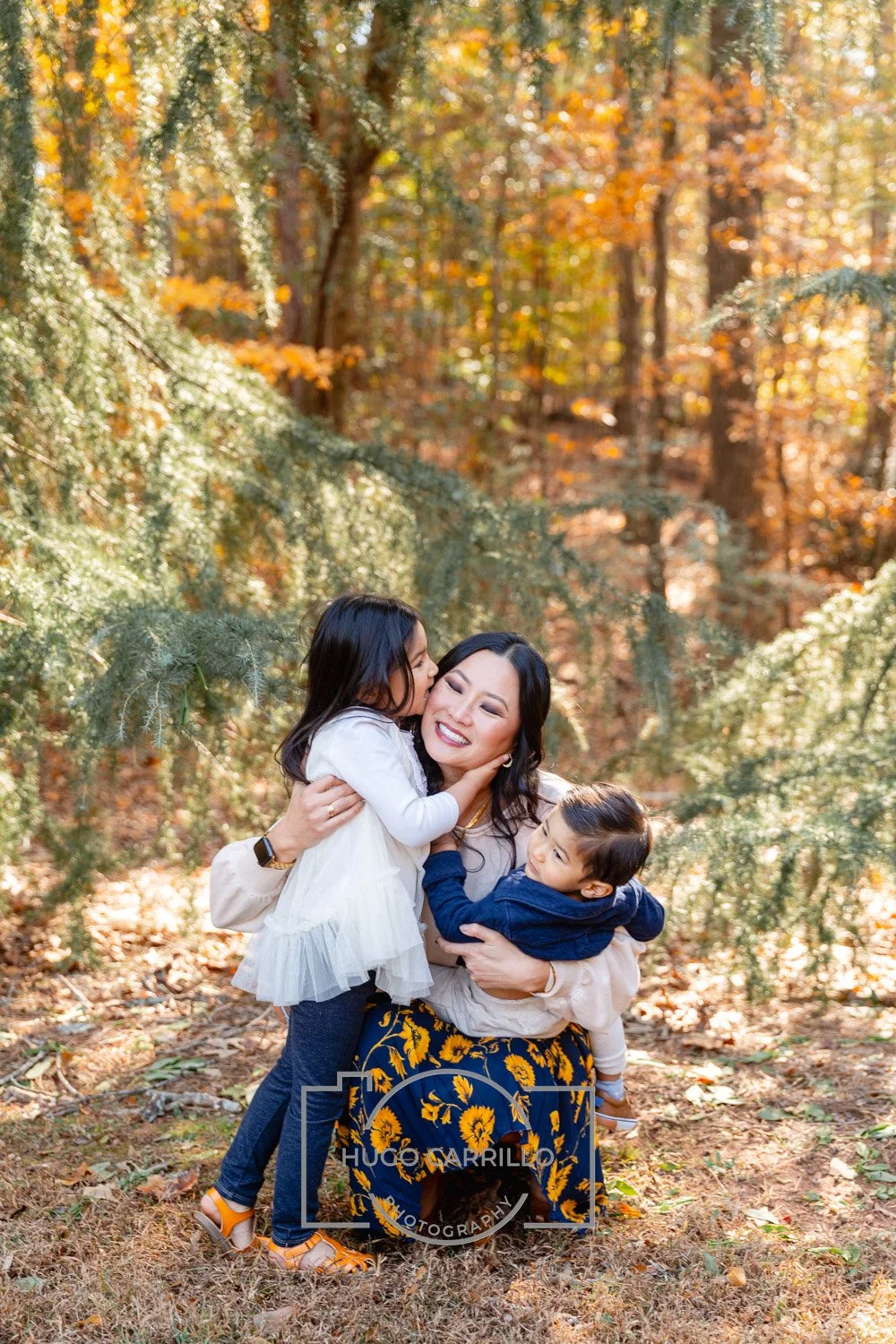 A woman with two children in an autumn forest, hugging and smiling, as the young girl kisses her on the cheek and the boy holds her from the side, with fall foliage and trees around.