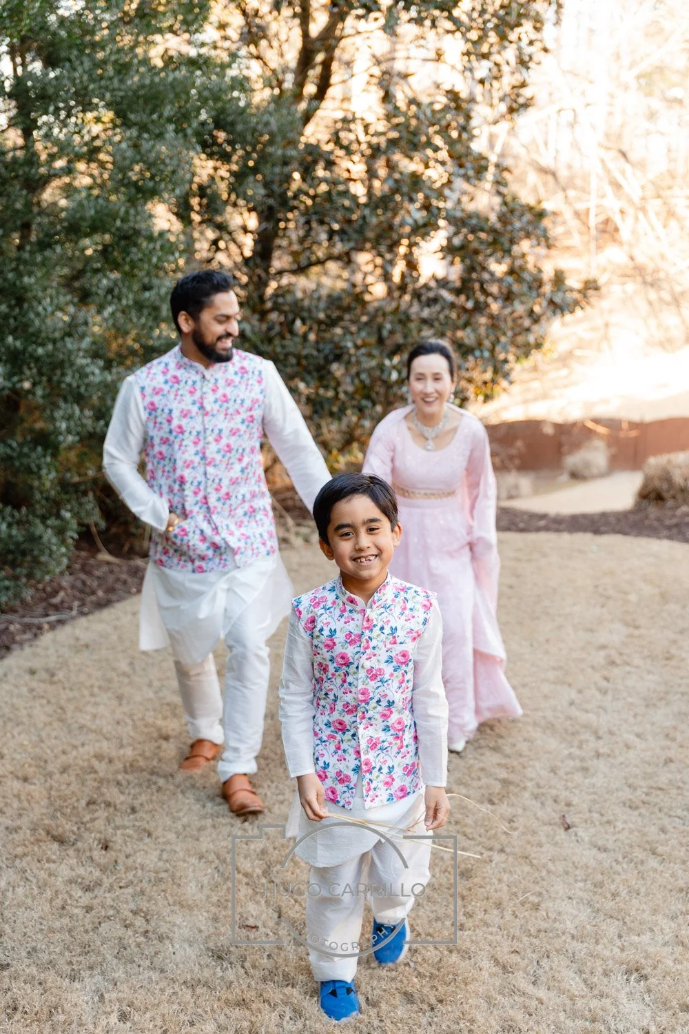 A happy family of three dressed in traditional Indian clothing walks outdoors on a sunny day. The young boy in the front is smiling, wearing a white kurta with a colorful floral vest, and holding a stick. Behind him are a man and a woman, both smilin