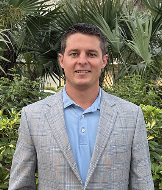 A man in a light gray checkered blazer and blue collared shirt stands outdoors in front of lush green tropical plants.