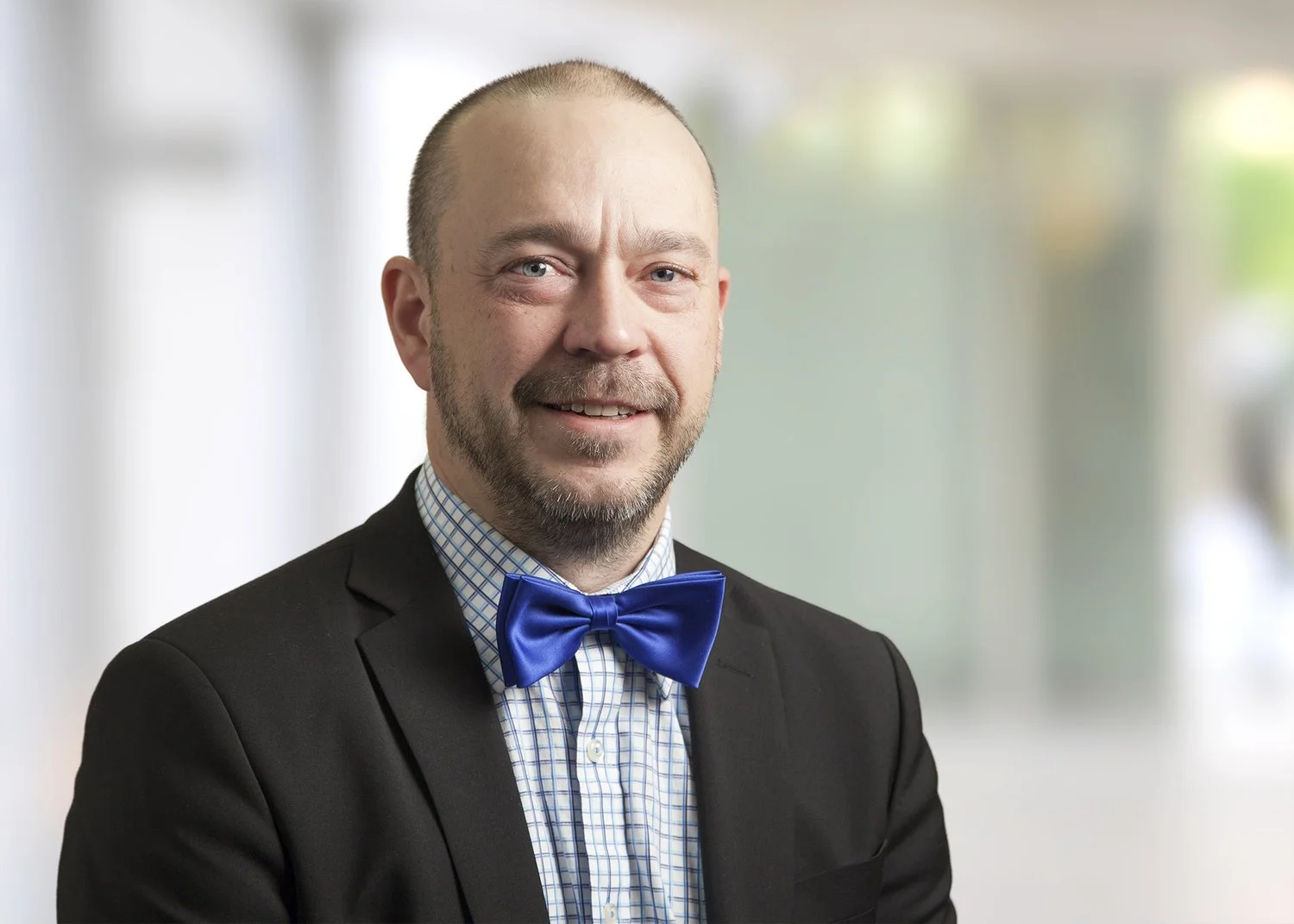 A man with a beard, wearing a black suit and a blue bow tie, smiling at the camera in a professional setting.