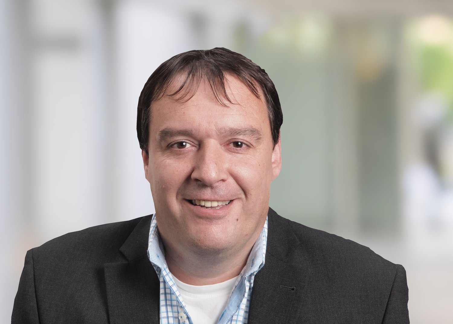 A smiling man wearing a dark suit jacket and a light blue checkered shirt in an office setting.