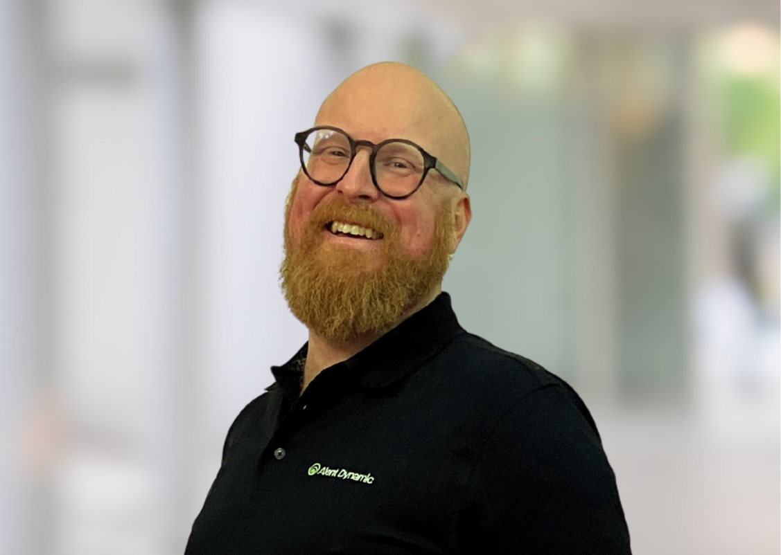 A man with a red beard and bald head wearing a dark suit with a white shirt and gold pocket square, standing with arms crossed in a blurred indoor setting.