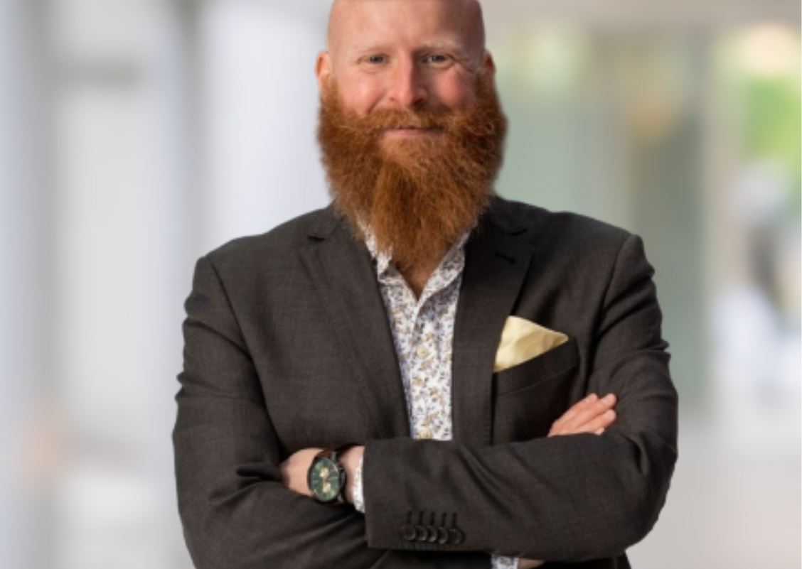 A man with a red beard and bald head wearing a dark suit with a white shirt and gold pocket square, standing with arms crossed in a blurred indoor setting.