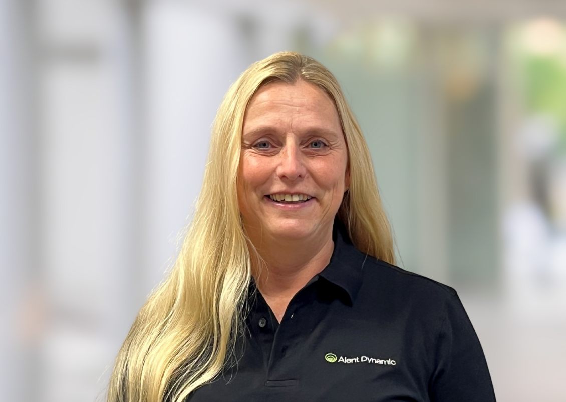 A woman with long blonde hair smiling, wearing a black polo shirt with a logo that says 'Alert Dynamic', standing indoors with a blurred background.