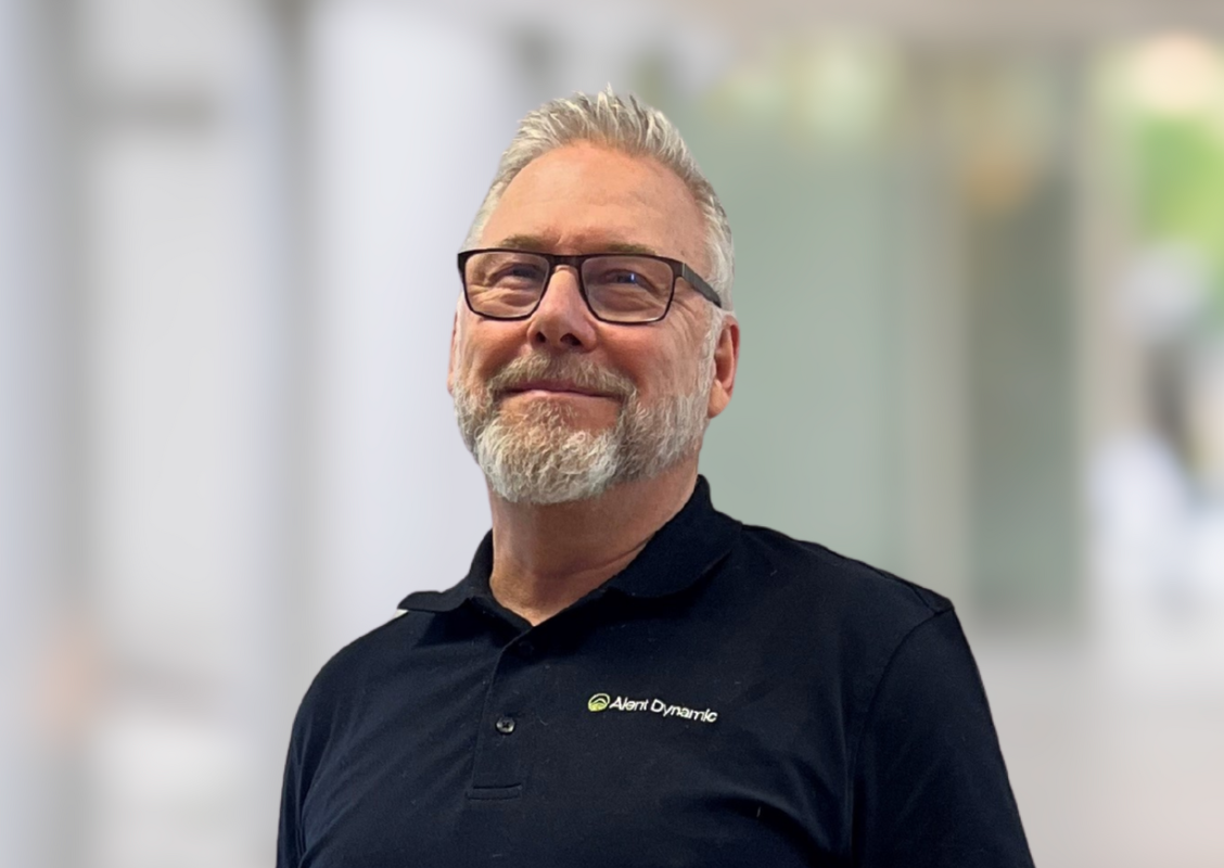 A middle-aged man with gray hair, beard, glasses, wearing a black polo shirt with the logo 'Alert Dynamic' on it, standing indoors near a blurred background.