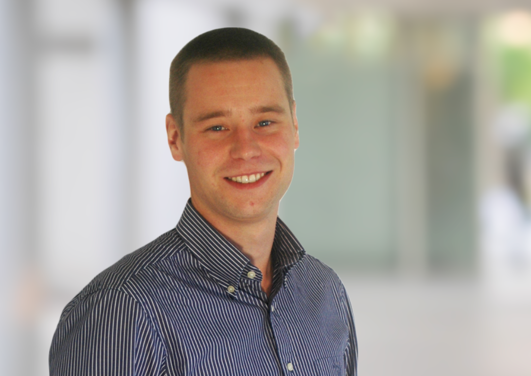 A young man with short dark hair, wearing a dark blue and white striped button-up shirt, smiling in an indoor setting with a blurred background.