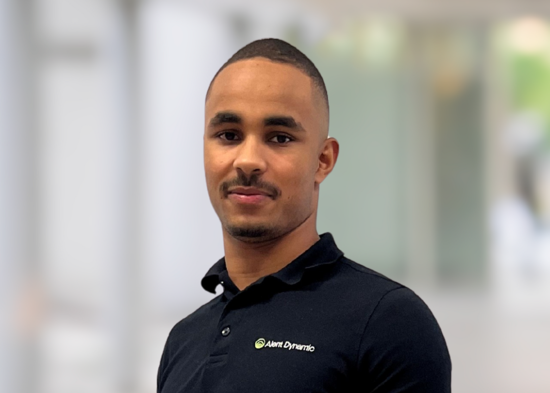 Young man with short hair and a goatee, wearing a black polo shirt with a logo on it, standing in a modern indoor setting.