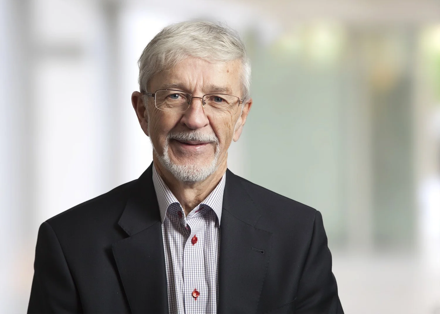 Portrait of an elderly man with white hair, glasses, and a beard, wearing a dark suit and a striped shirt, smiling in a professional setting.