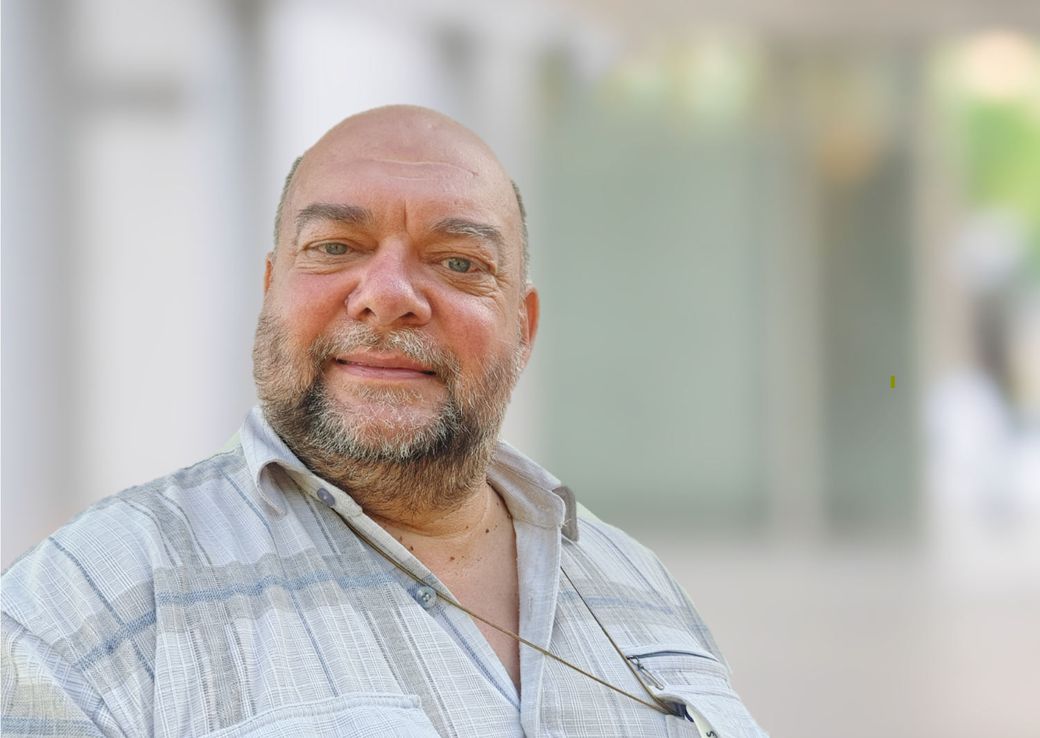 A middle-aged man with a beard and short hair smiling and looking at the camera, wearing a light plaid shirt, outdoors with a blurred background.