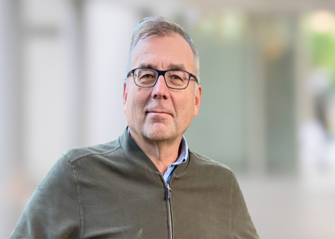 A middle-aged man with short gray hair and glasses, wearing a dark zip-up jacket and a collared shirt, standing indoors with a blurred background.