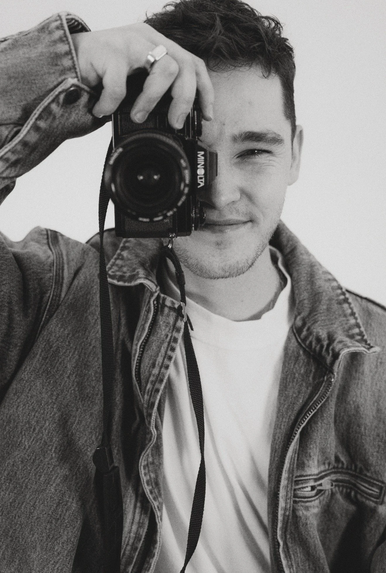A young man with short dark hair and a slight beard, smiling slightly, taking a black and white selfie with a vintage camera. He is wearing a denim jacket over a white shirt, standing against a plain background.