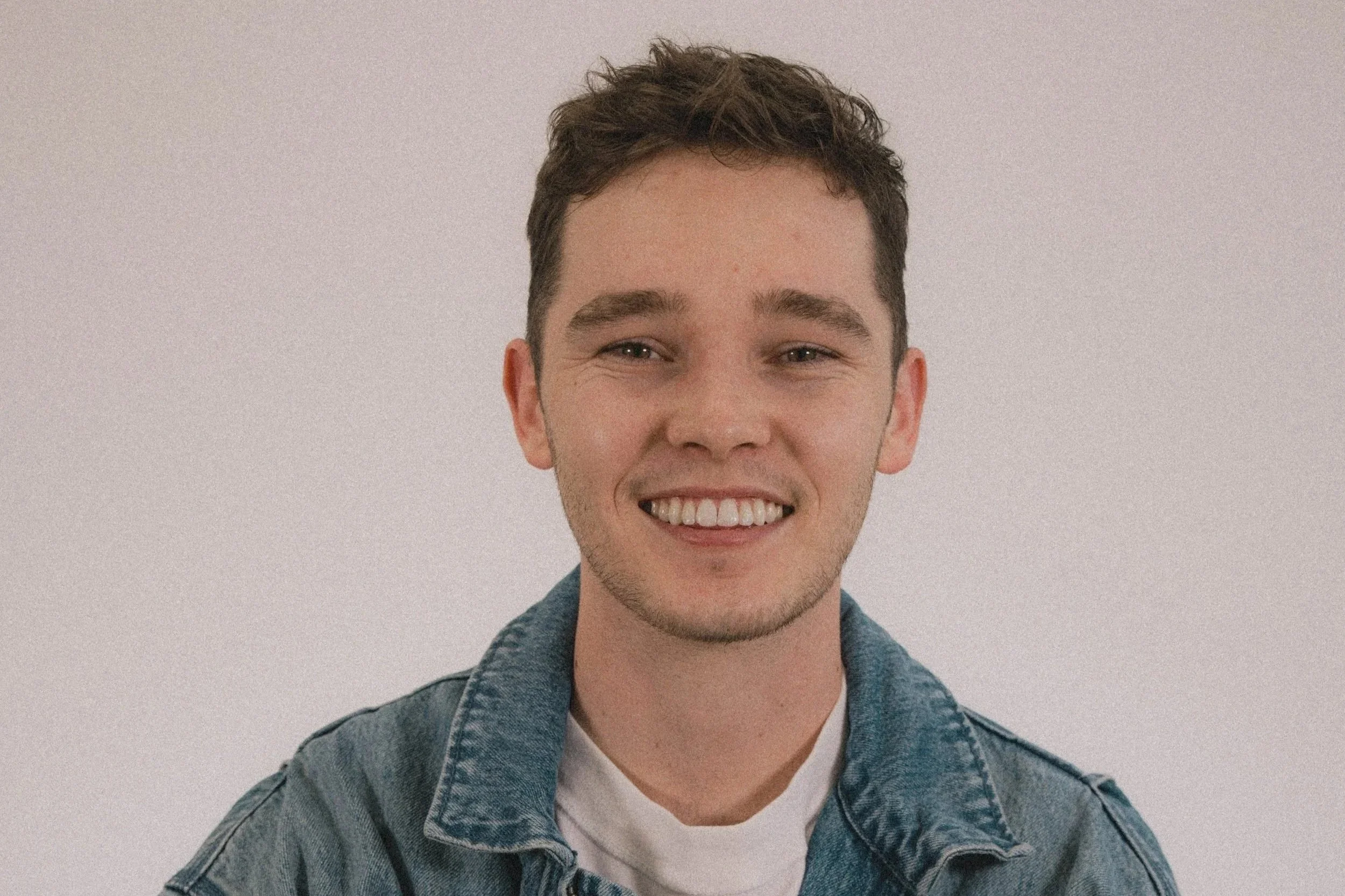 A young man with short, curly brown hair, smiling with visible teeth, wearing a denim jacket over a white t-shirt, against a plain light-colored background.