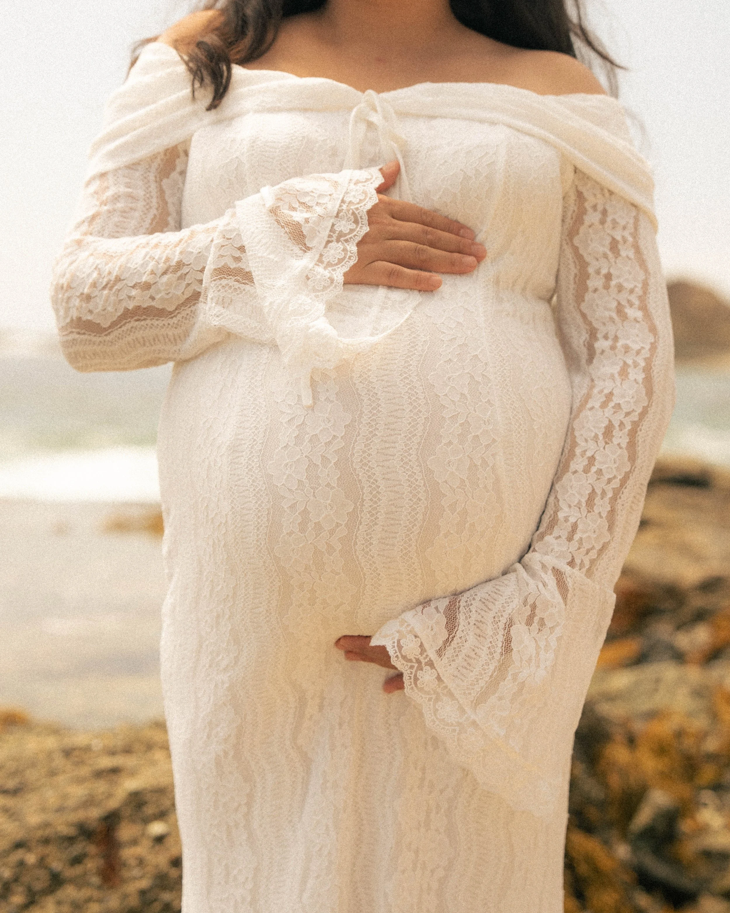 A woman wearing a white lace off-shoulder dress with long sleeves and ruffled cuffs, holding her hand over her chest, standing outdoors in a natural setting.