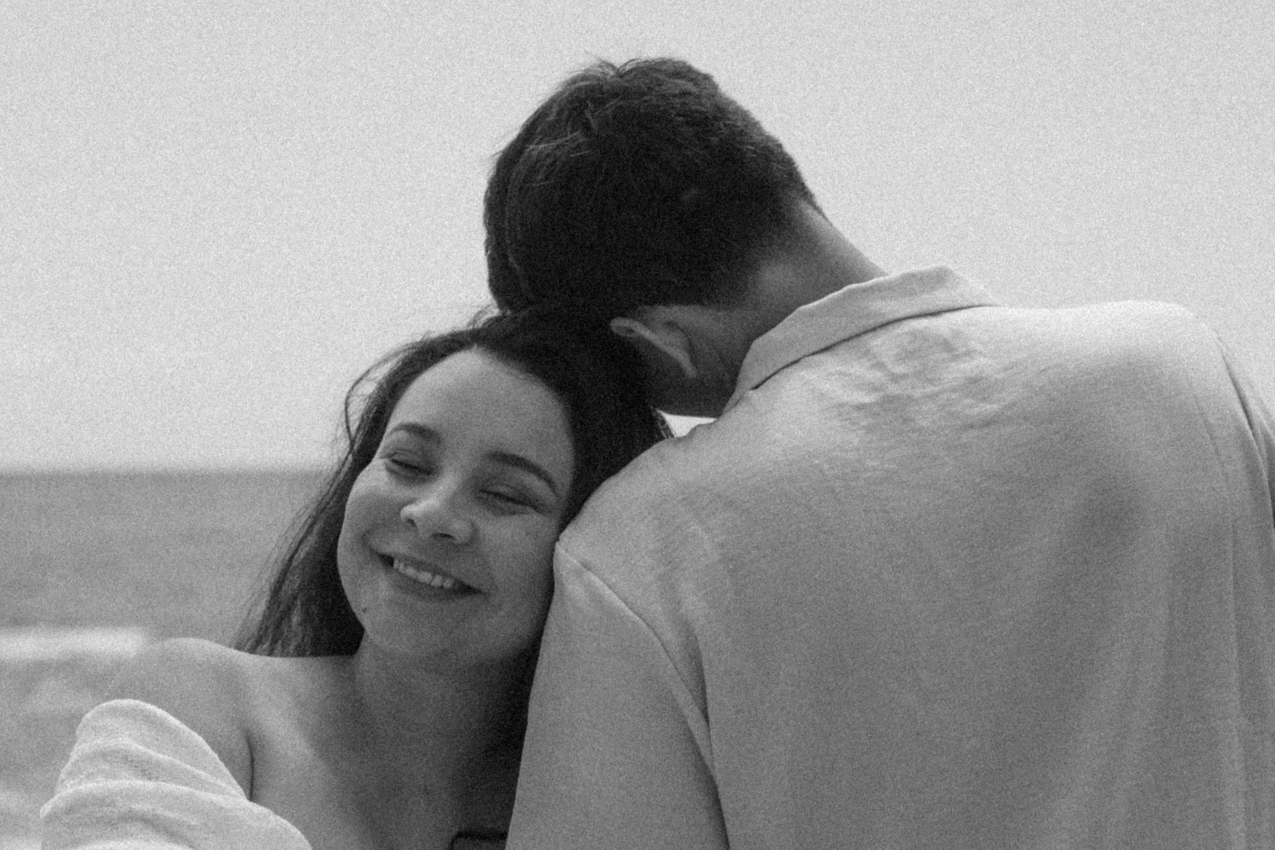 A woman with long dark hair smiling with closed eyes, embracing a man with short dark hair in an affectionate hug.