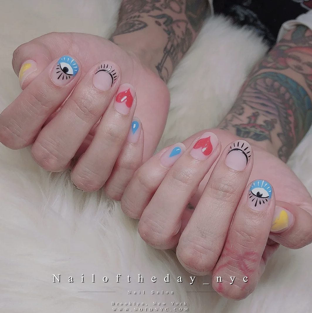 Close-up of a person with colorful, artistic nail designs, featuring eye and abstract patterns, on both hands, with visible tattoos on arms, resting on a white furry surface.