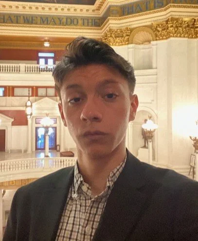A man wearing glasses and a navy suit with a pink shirt and patterned tie, set against a blue background.