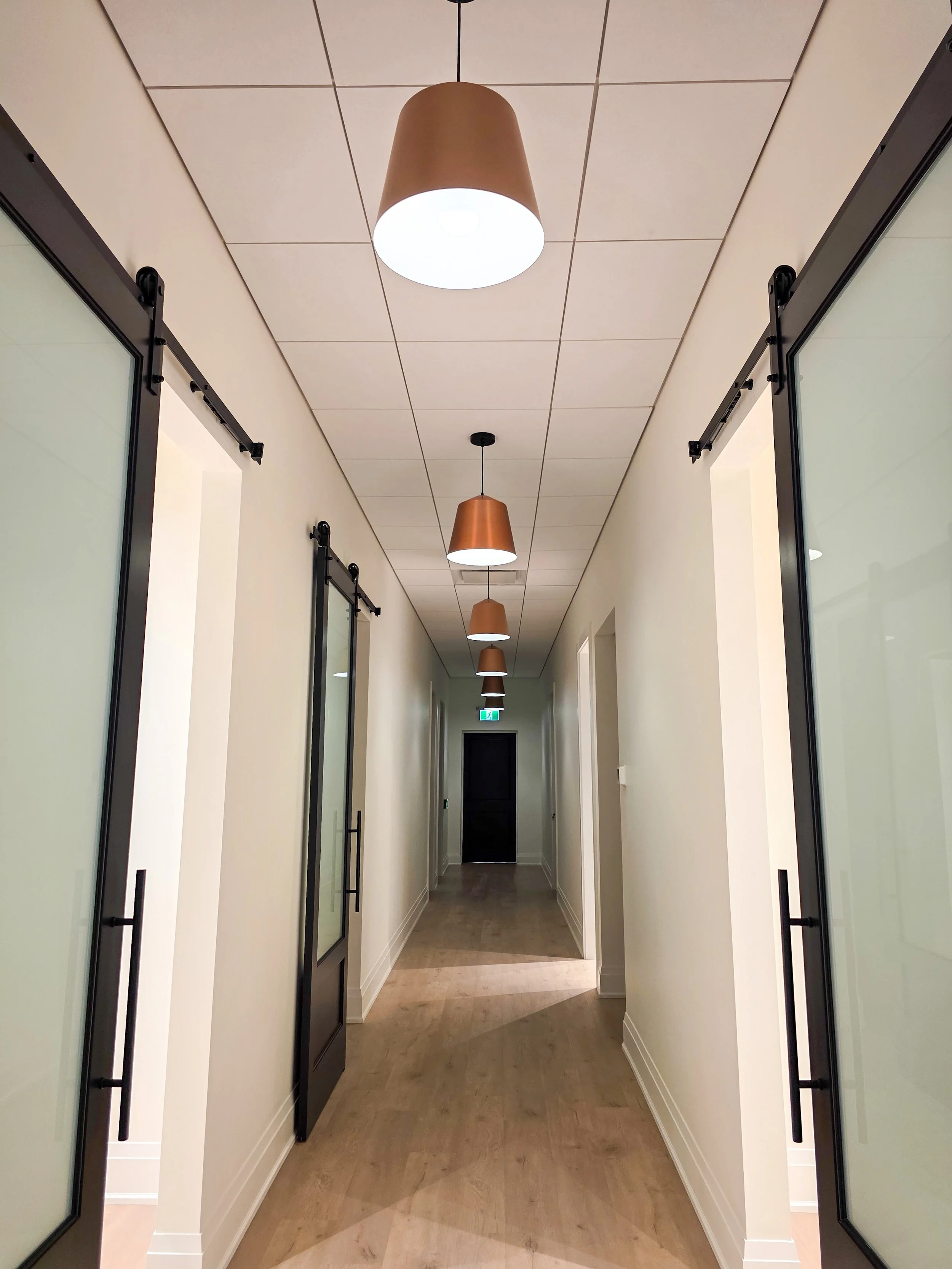 A hallway with a series of hanging pendant lights, white walls, glass-paneled doors with black handles and sliding hardware, and a light-colored wooden floor.