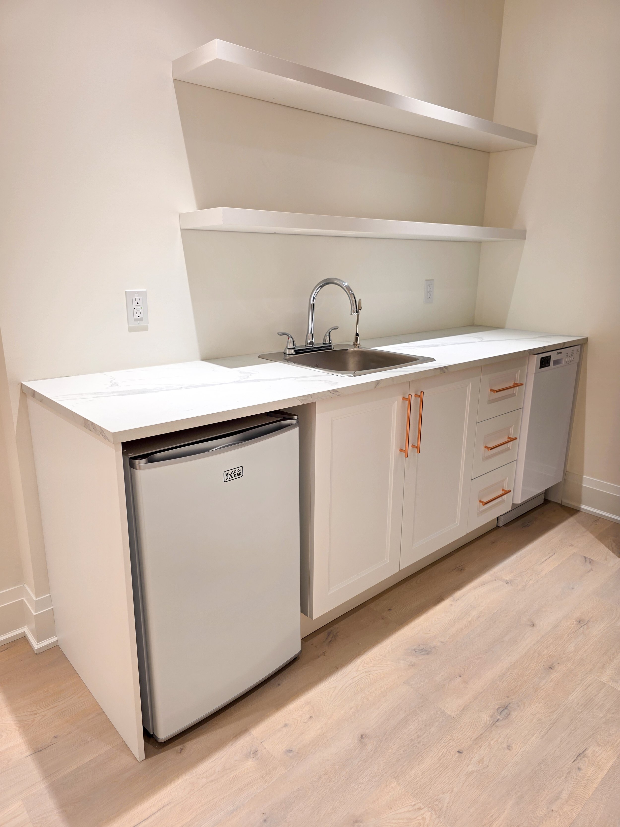 A modern kitchen area with white cabinetry, a small refrigerator, a sink with a chrome faucet, and two open white shelves on a white wall, with light wood flooring.