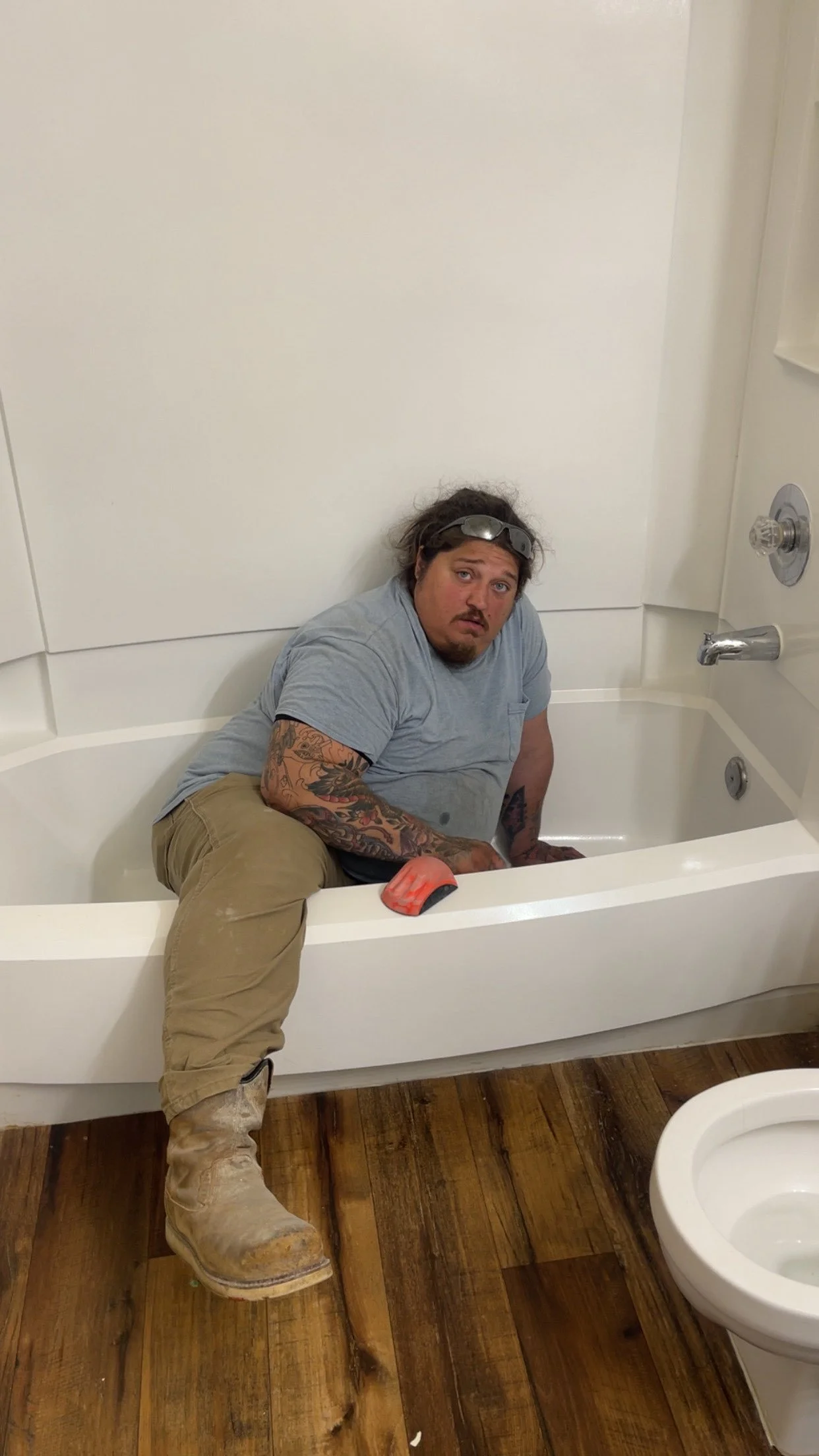 A man with tattoos, wearing a gray shirt, beige pants, and worn-out boots, sitting in a bathtub with a surprised or concerned expression on his face, in a bathroom with wooden flooring and a white toilet nearby.