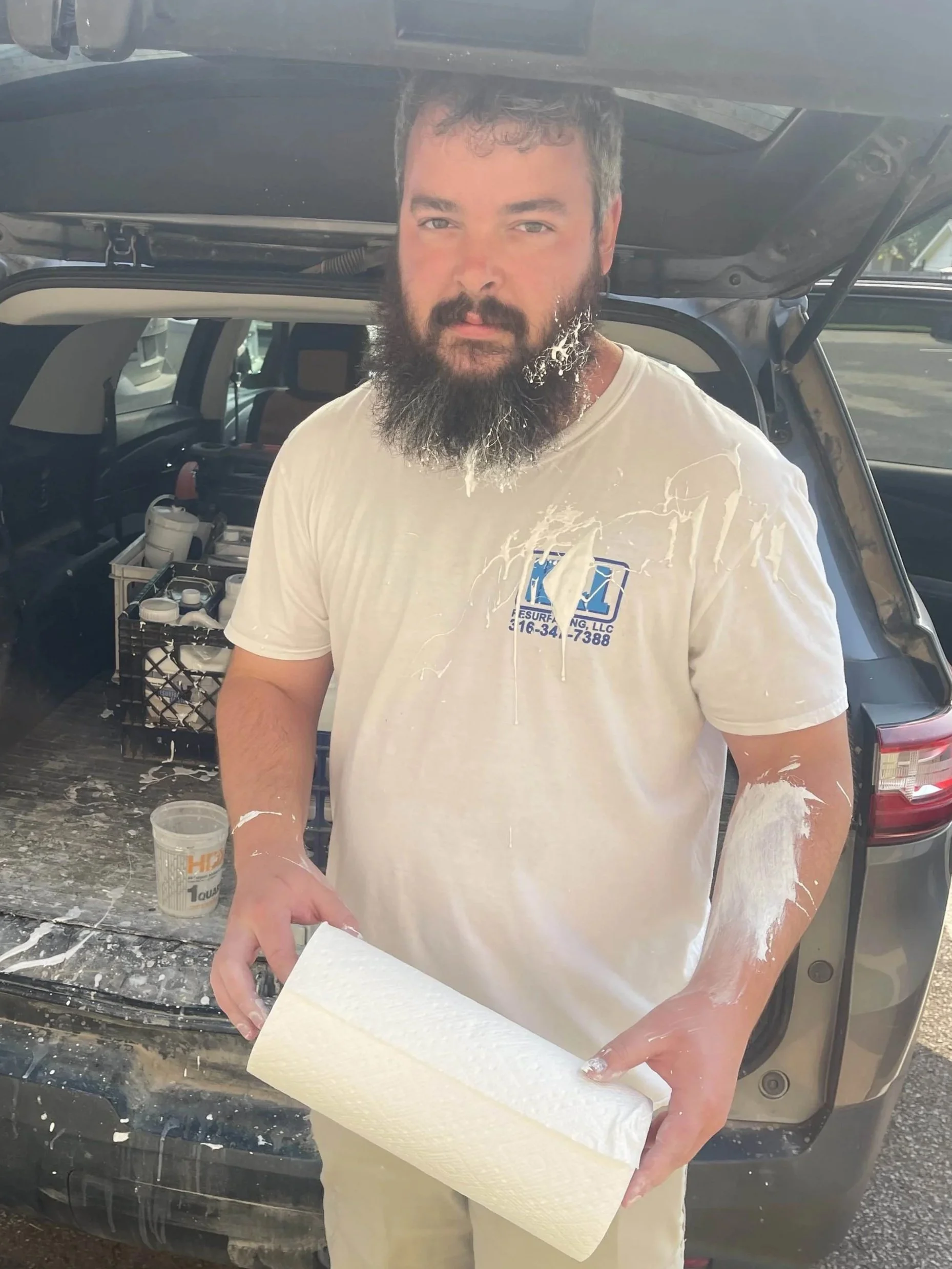 A bearded man standing in front of an open car trunk, holding a roll of paper towels. He is wearing a white t-shirt with paint splatters and has paint on his arms and shirt. There are paint supplies in the car trunk.