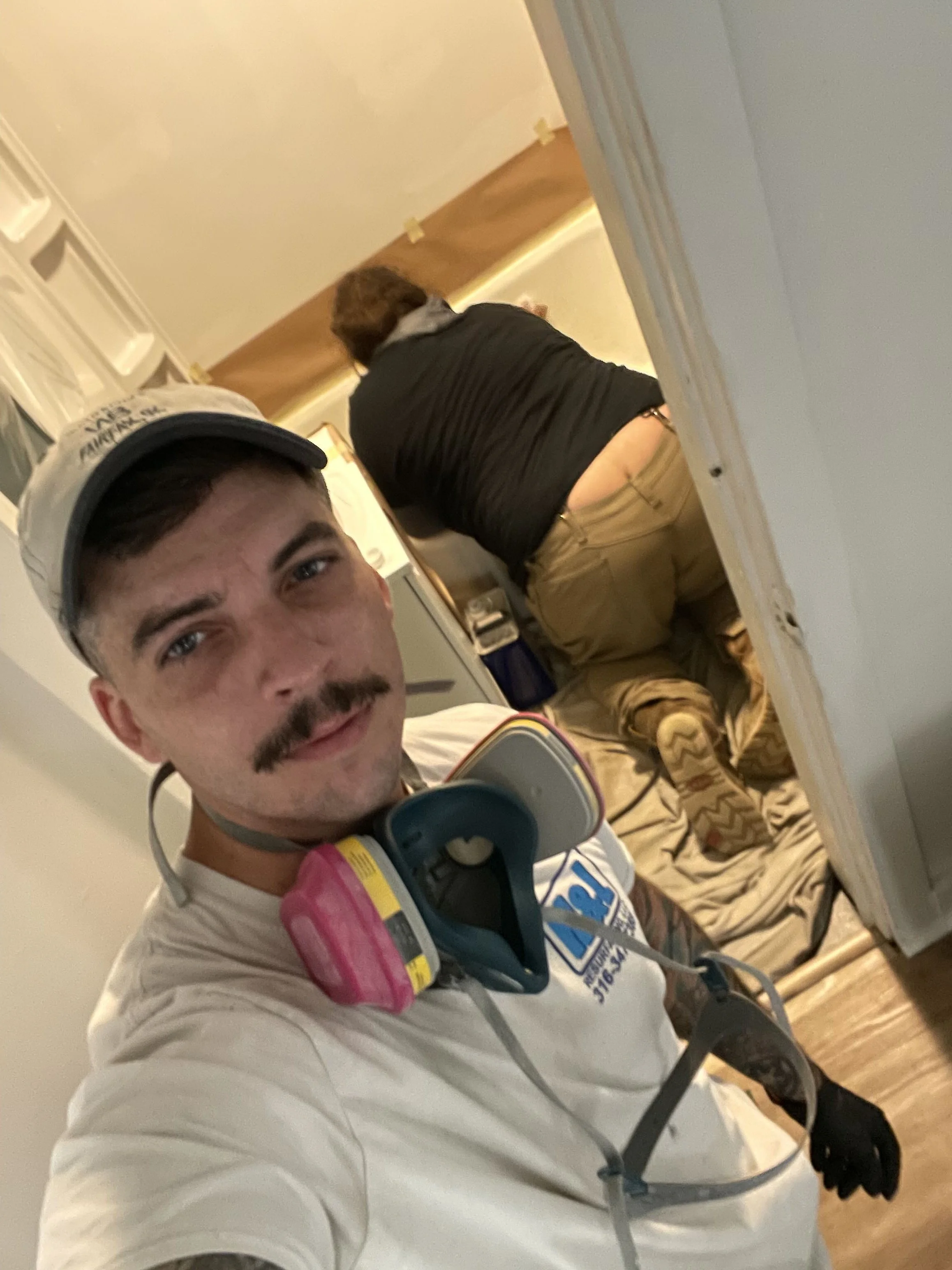 A young man taking a selfie while working on a home improvement project. He has a mustache, a cap, and safety equipment around his neck. In the background, a woman with long hair is kneeling on the floor, working near a doorway.