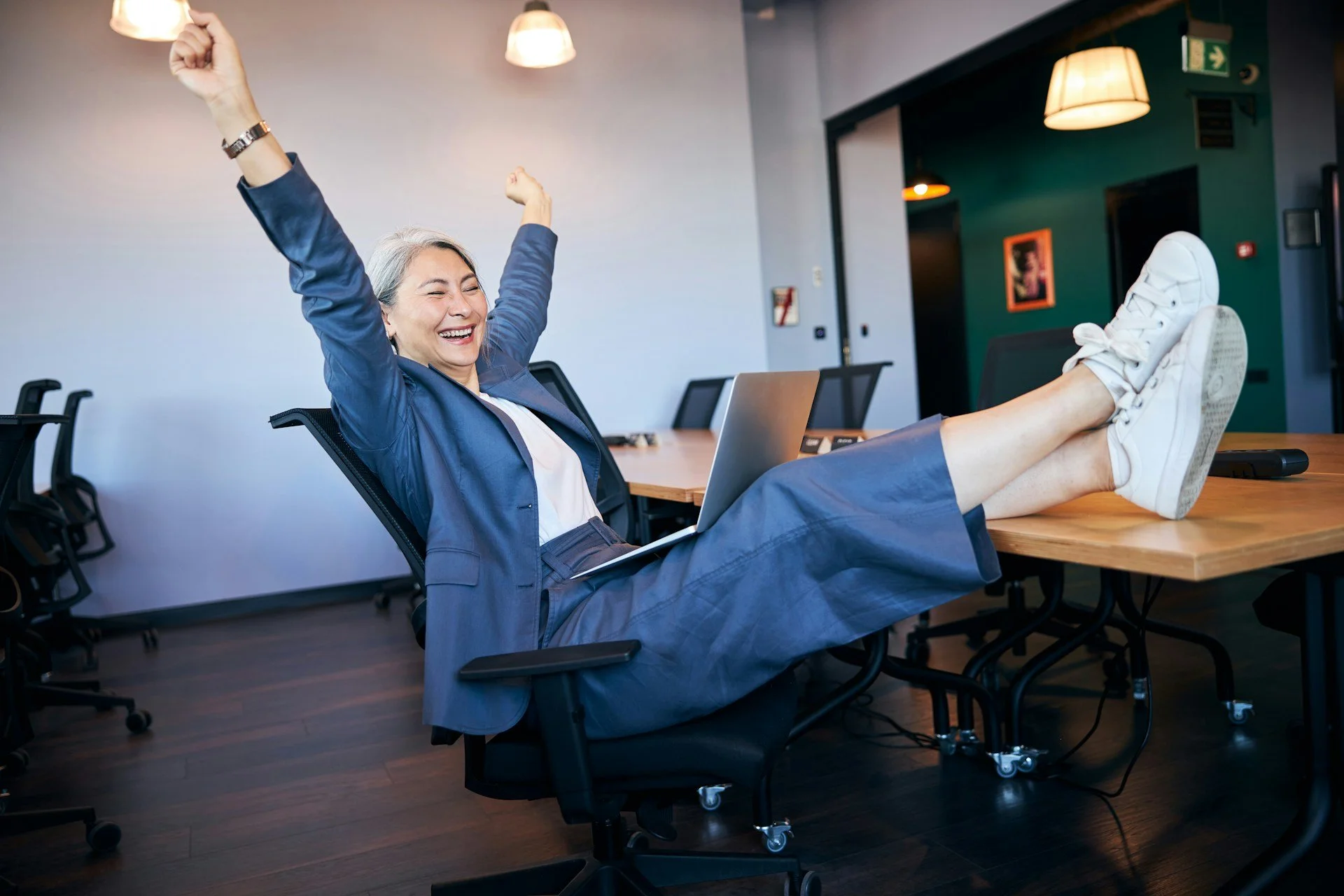 Joyful businesswoman with laptop raising hands and smiling while sitting in chair and placing her feet on desk