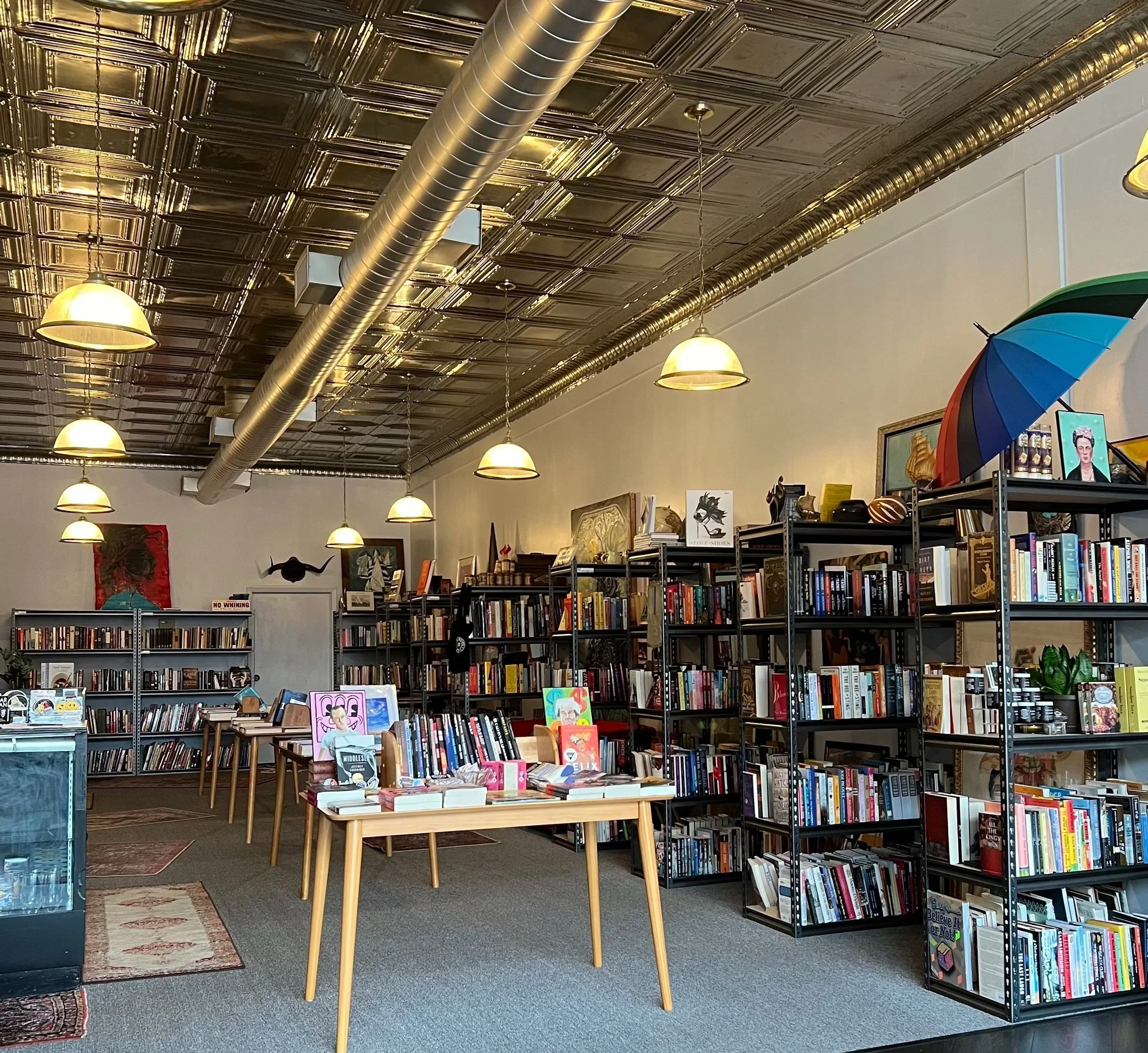 Photograph of the interior of a bookstore with bookshelves filled with books, hanging pendant lights, a patterned rug, and a decorative ceiling with metallic panels and a large metal air vent.