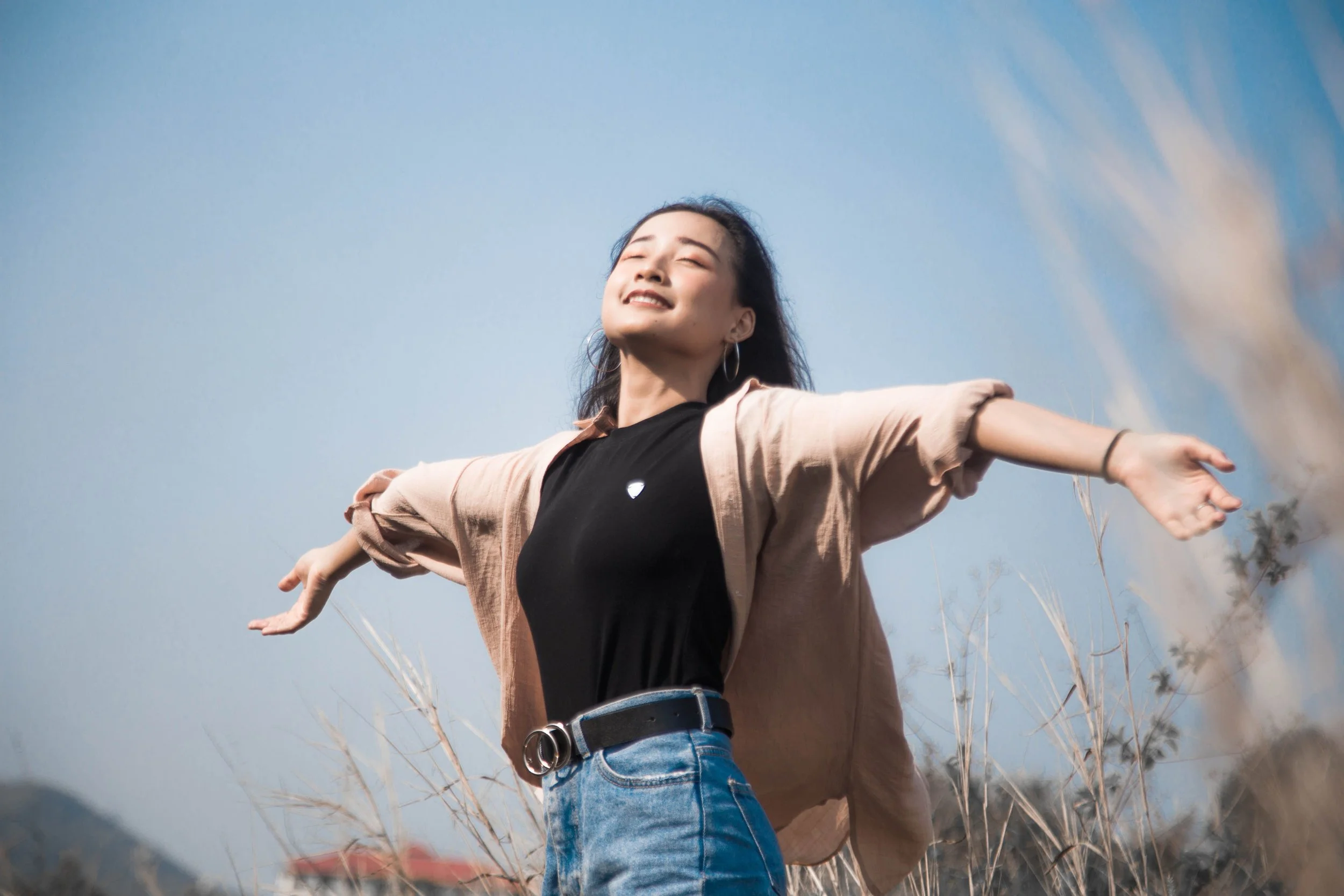 A woman with long dark hair and hoop earrings happily smiling with arms outstretched in a field of tall, dry grass during daytime, with blue sky overhead.