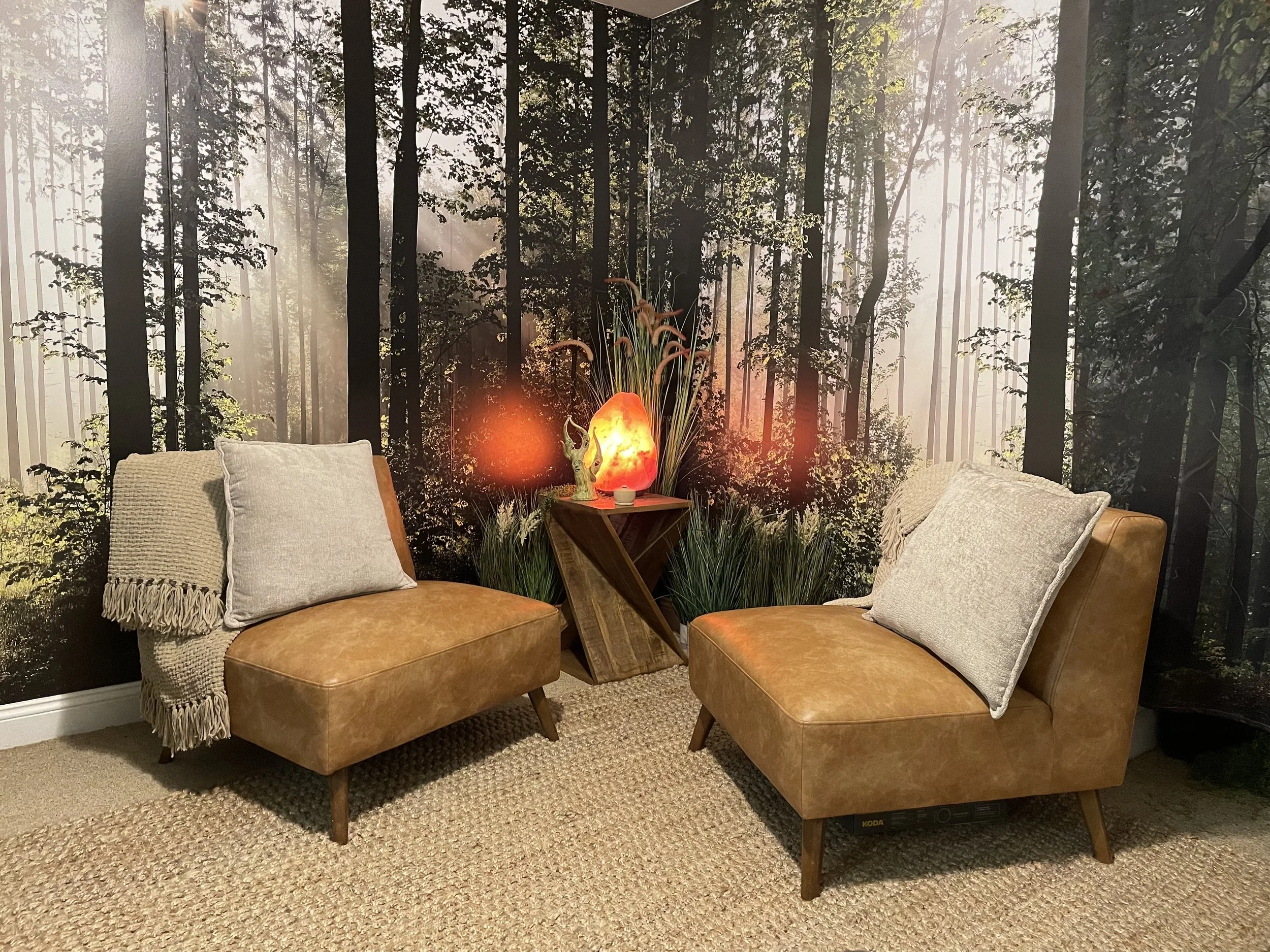 An image of Dr. Zeus's office with a wall mural of a dense forest. There are two tan leather sofas with off-white and gray throw pillows, a small wooden table with a salt lamp, and a beige rug on a carpeted floor.