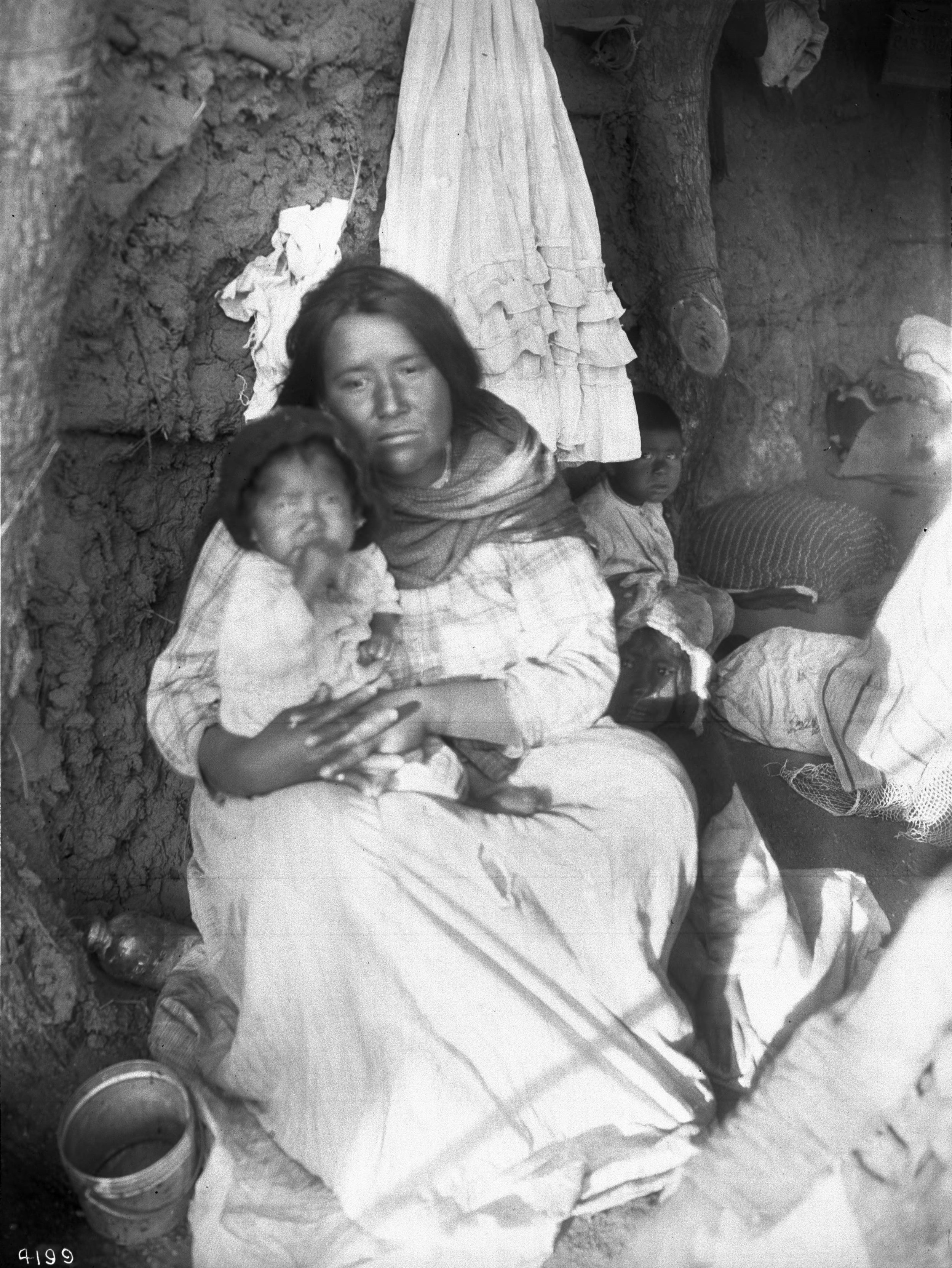 A black and white photo of a woman and her three children sitting together, both looking at the camera. They are sitting against a mud wall, with a cloth hanging above them and a plastic bag and a bucket nearby.