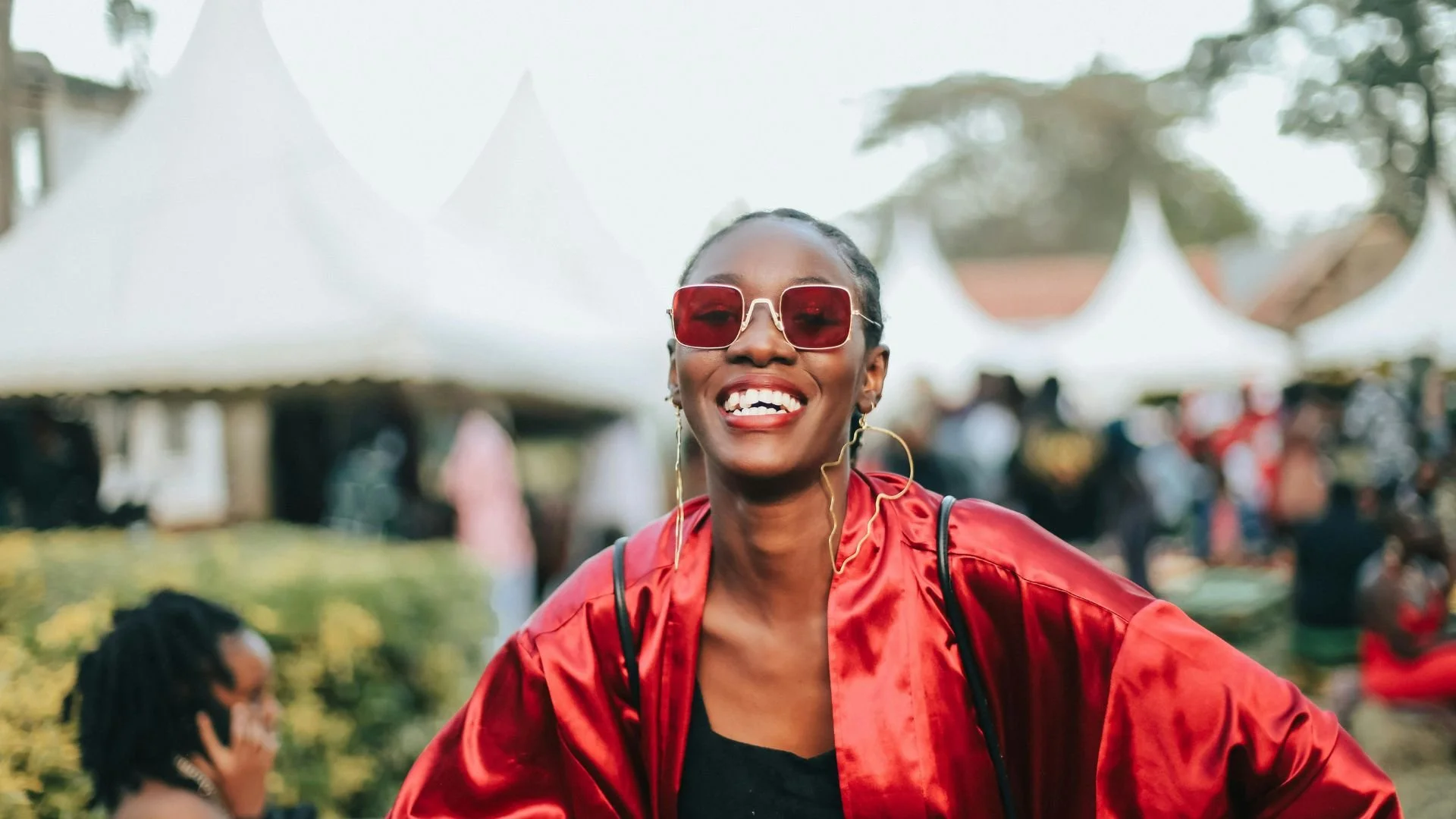 Lady smiling, wearing a red shirt and red sunglasses, large gold earrings. She is at a daytime outdoor event. White tents are in the background. The weather is great, and so are the vibes!