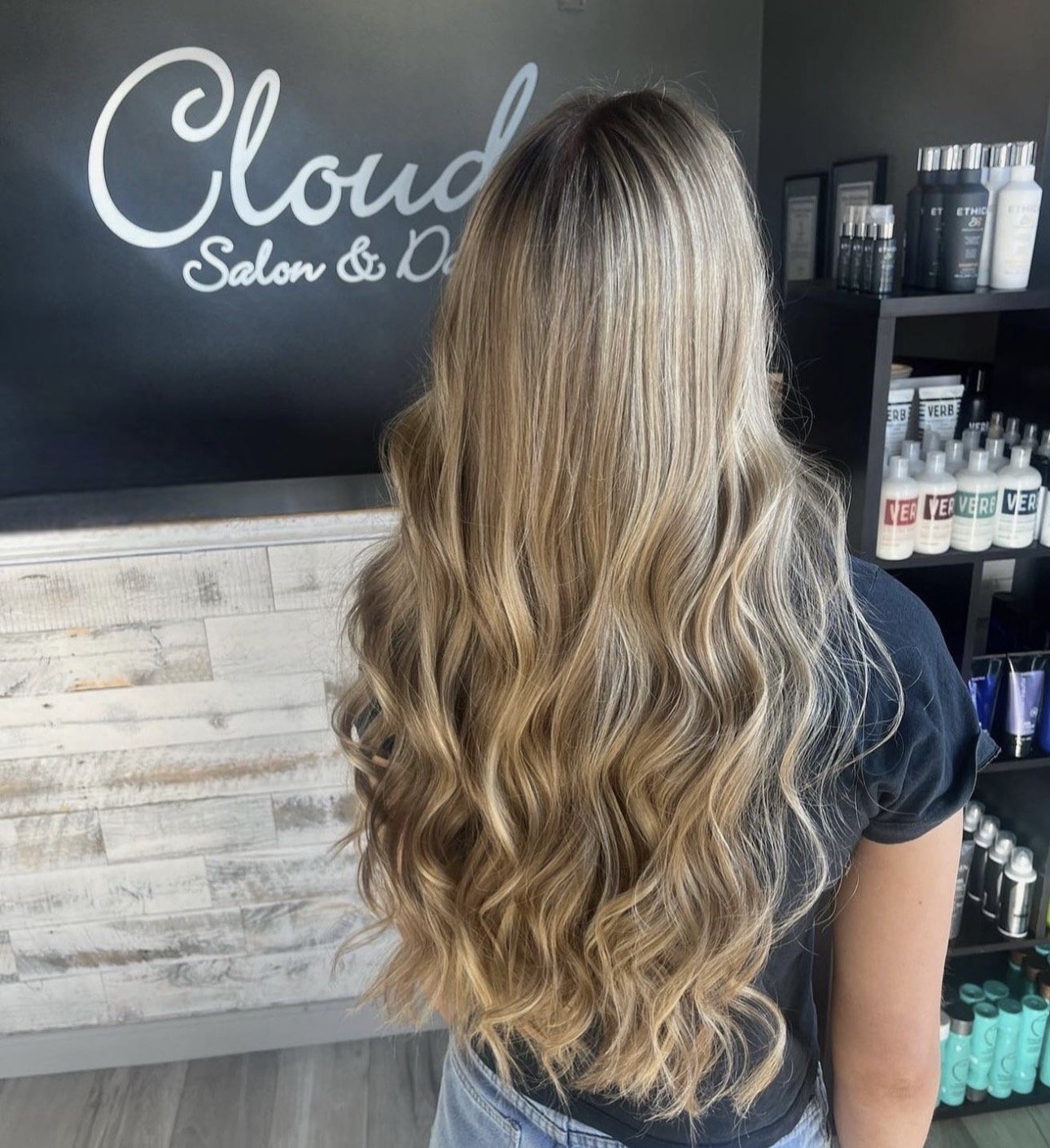 A woman with long, wavy blonde hair in a salon, standing in front of a black wall with the words "Cloud Salon &".