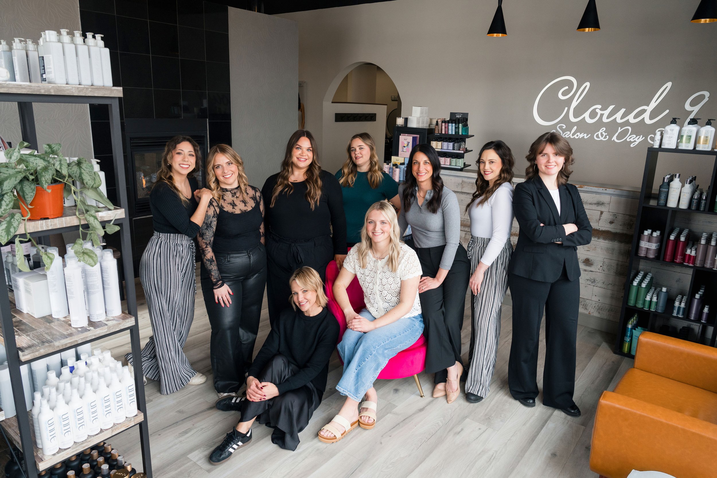 Group of women posing inside a salon with shelves of hair and skincare products, some sitting on a pink chair, others standing, and a wall with the text 'Cloud 9 Salon & Day Spa'.
