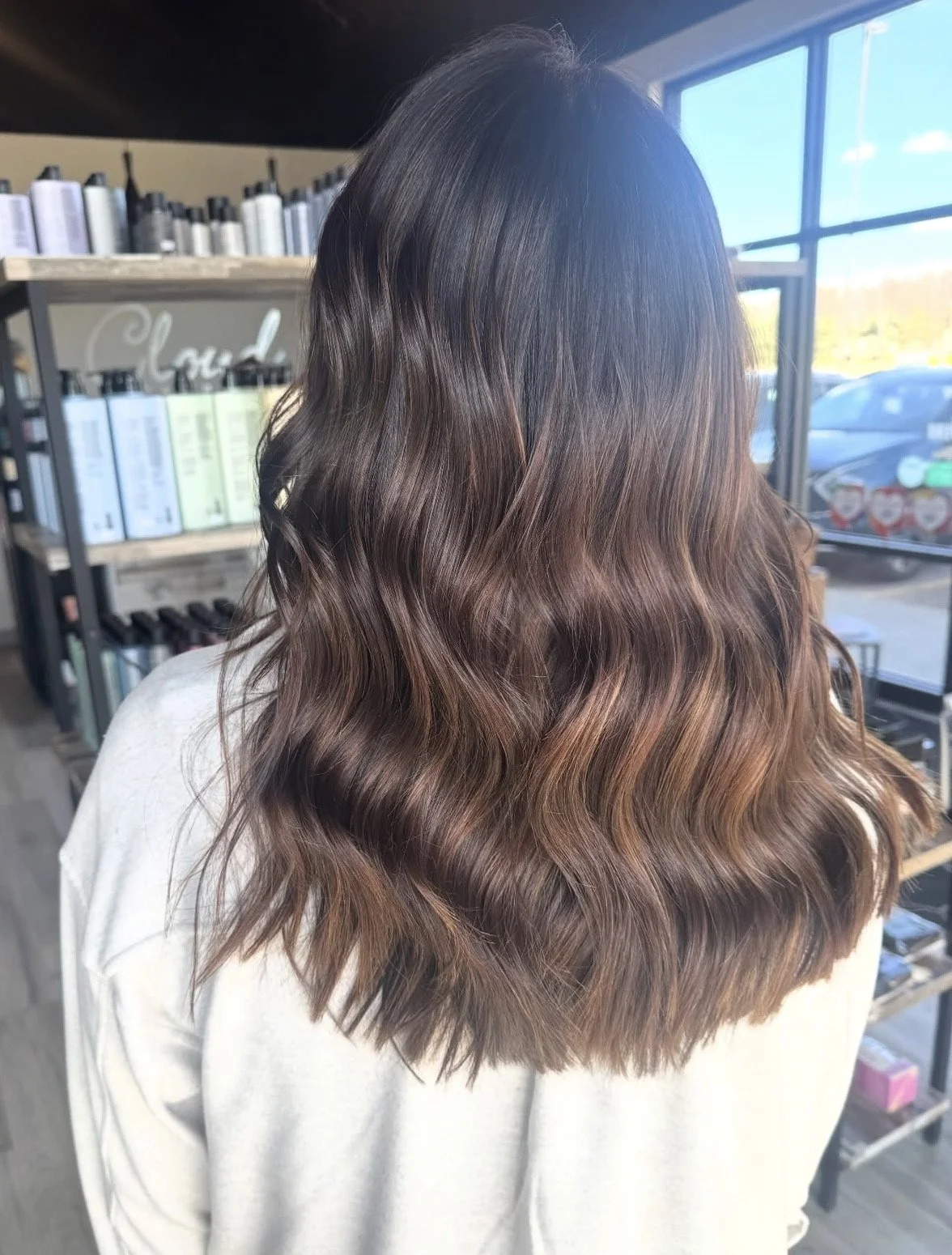 Back of a woman with long, wavy hair that has a gradient from dark brown at the roots to caramel at the ends, inside a hair salon.
