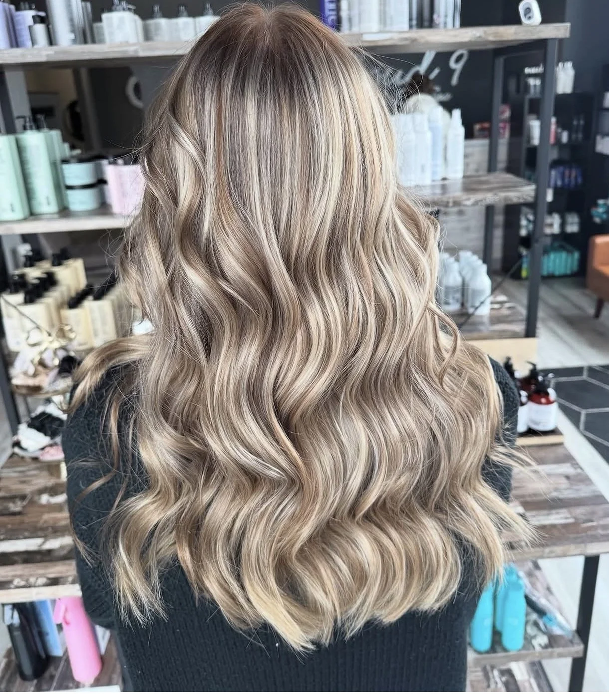 A woman with long, wavy blonde hair sitting in a salon with shelves of hair products in the background.