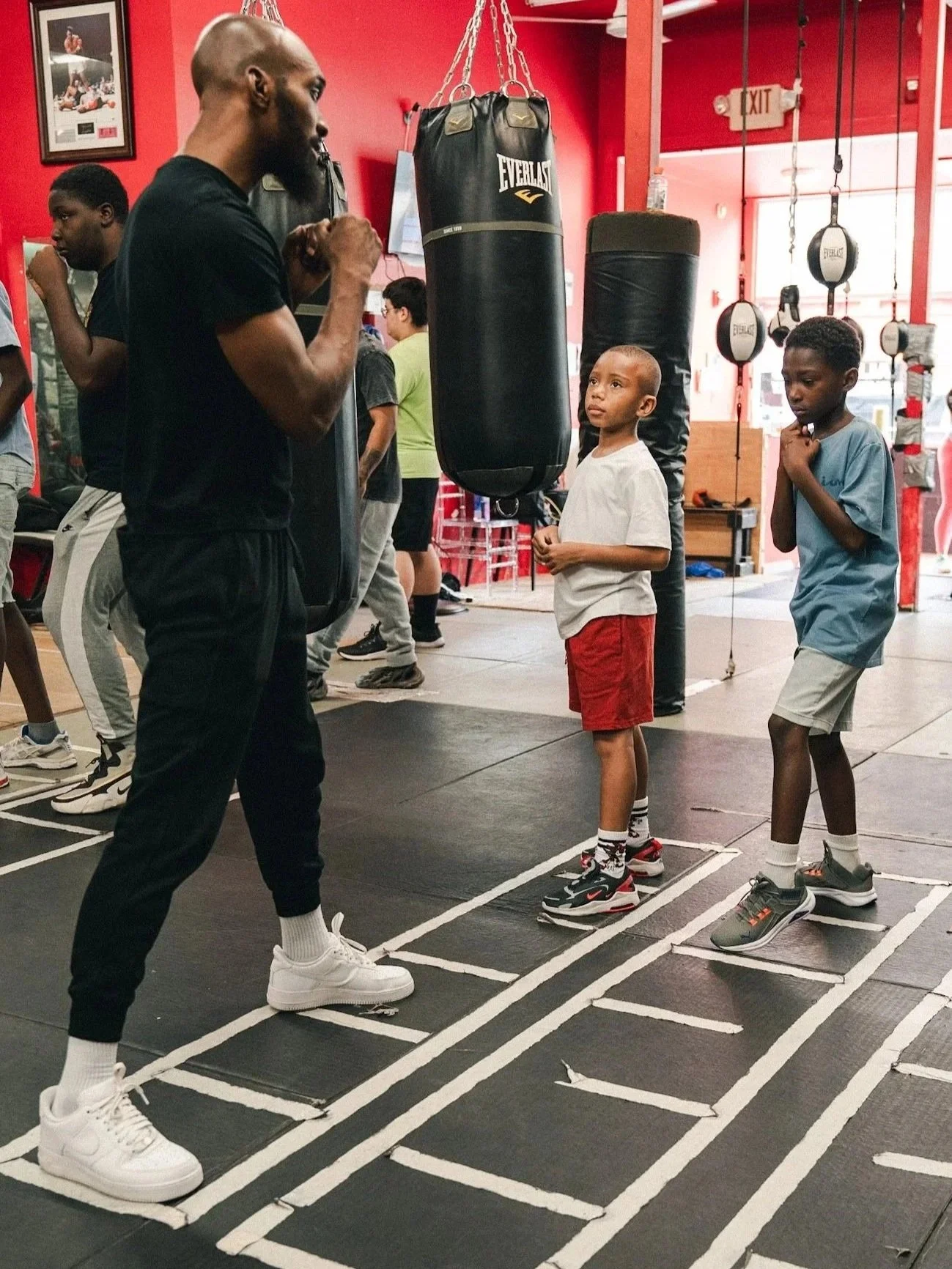 A trainer giving a boxing lesson after school to two young boys at a gym with punching bags and other training equipment.
