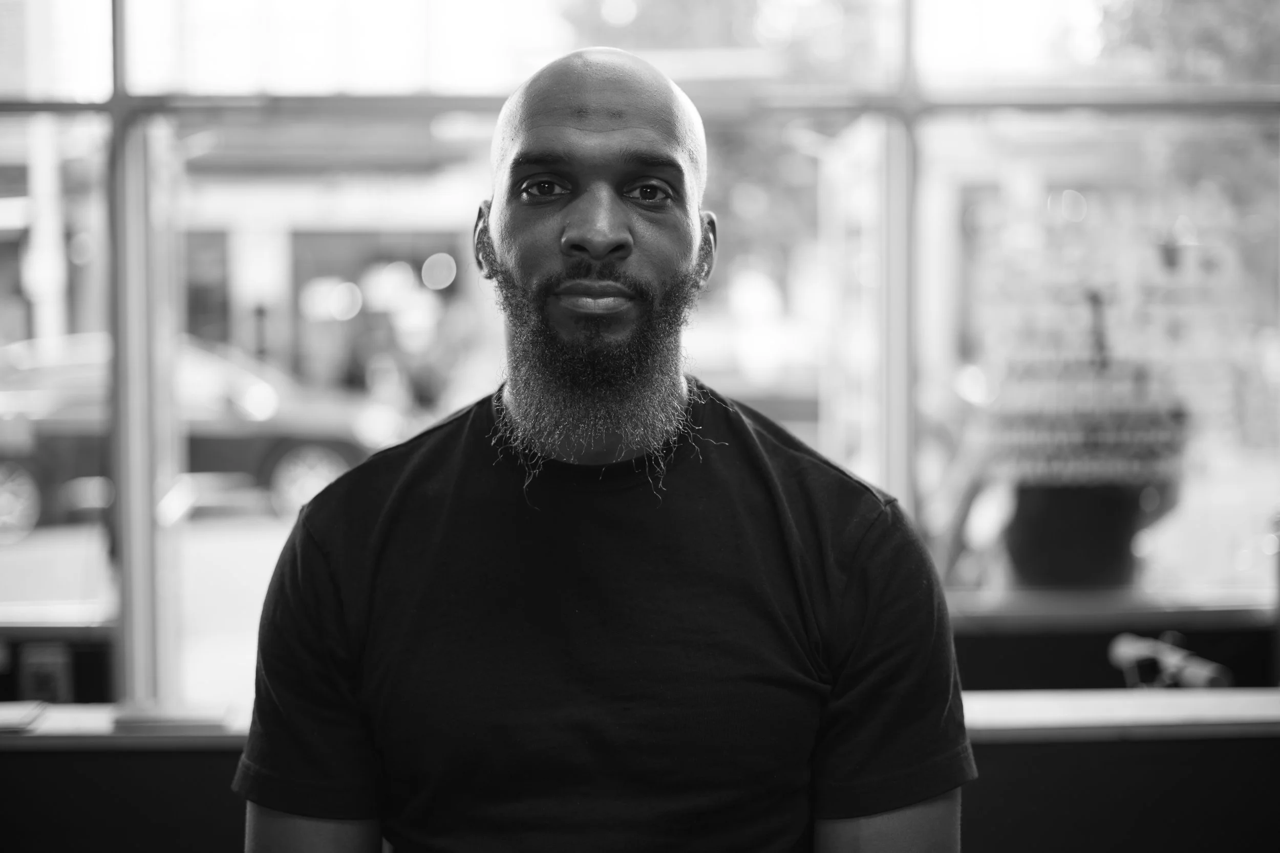 Black and white photo of a bald man with a beard sitting indoors with large windows behind him, showing cars outside.