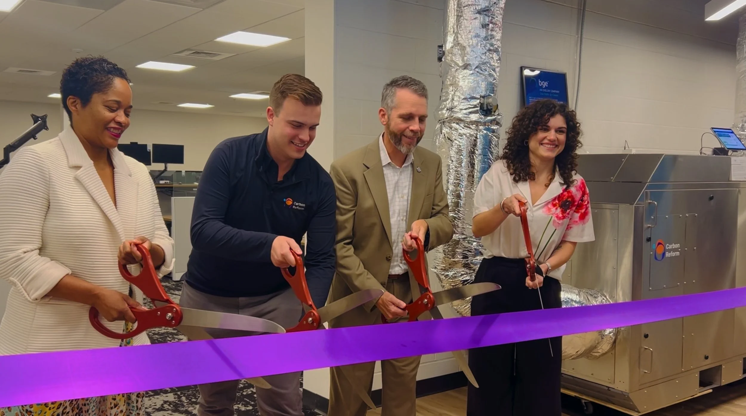 Four people participating in a ribbon-cutting ceremony with scissors, smiling, in a modern industrial or office setting.