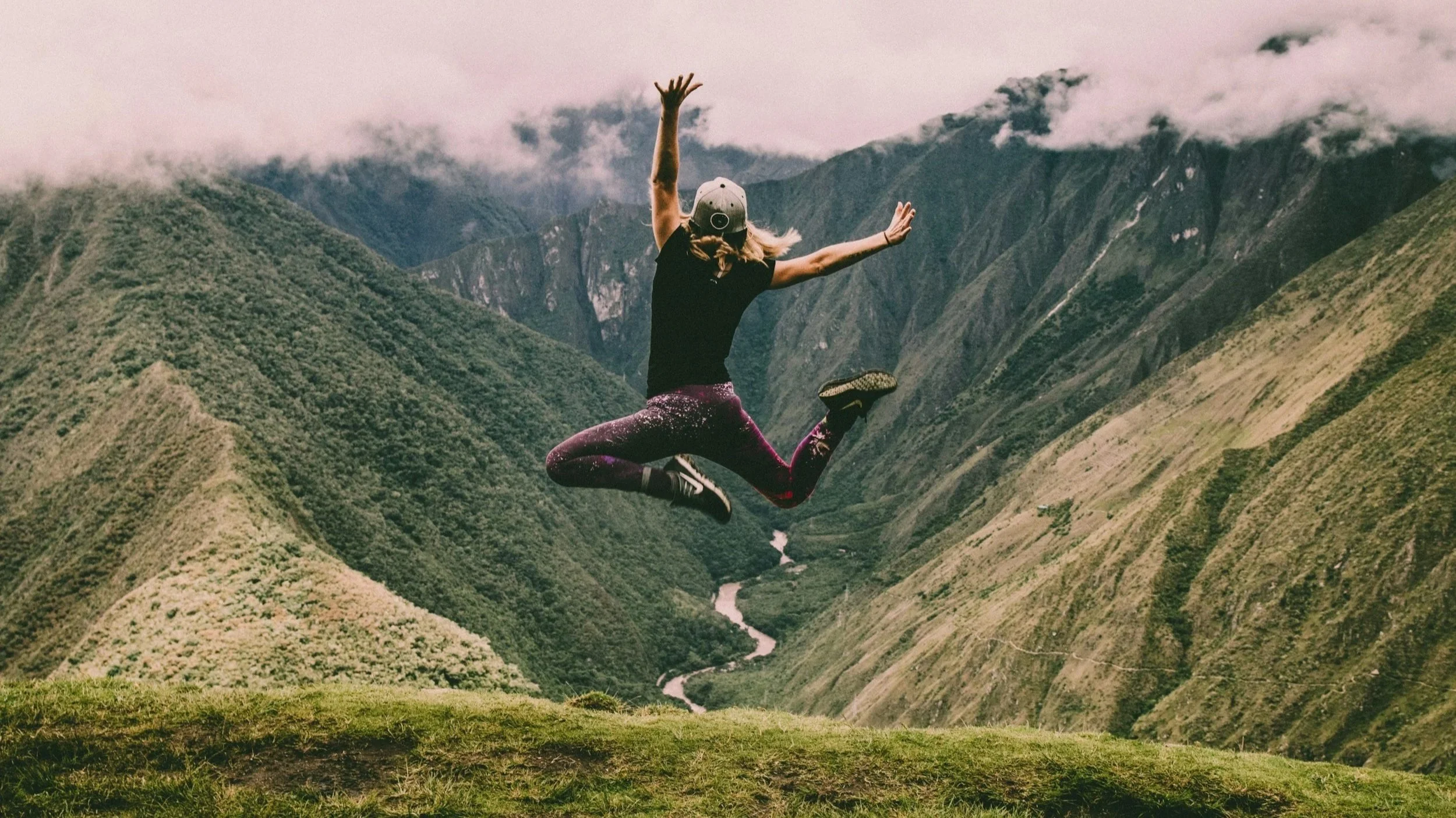 Mujer vestida con un pantalón y camiseta y gorra brincando de alegría mientras contempla un paisaje frente a ella de montañas verdes en donde abajo se ve cómo está corriendo un río. Ella decidió conscientemente disfrutar del momento presente