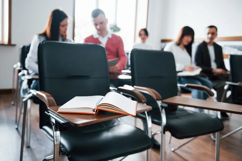 Sala de clases con varias sillas y un libro abierto en una de ellas, y estudiantes en el fondo reflexionando y conversando acerca de los cursos de Semiología de la Vida Cotidiana, cruciales para transformar su vida.