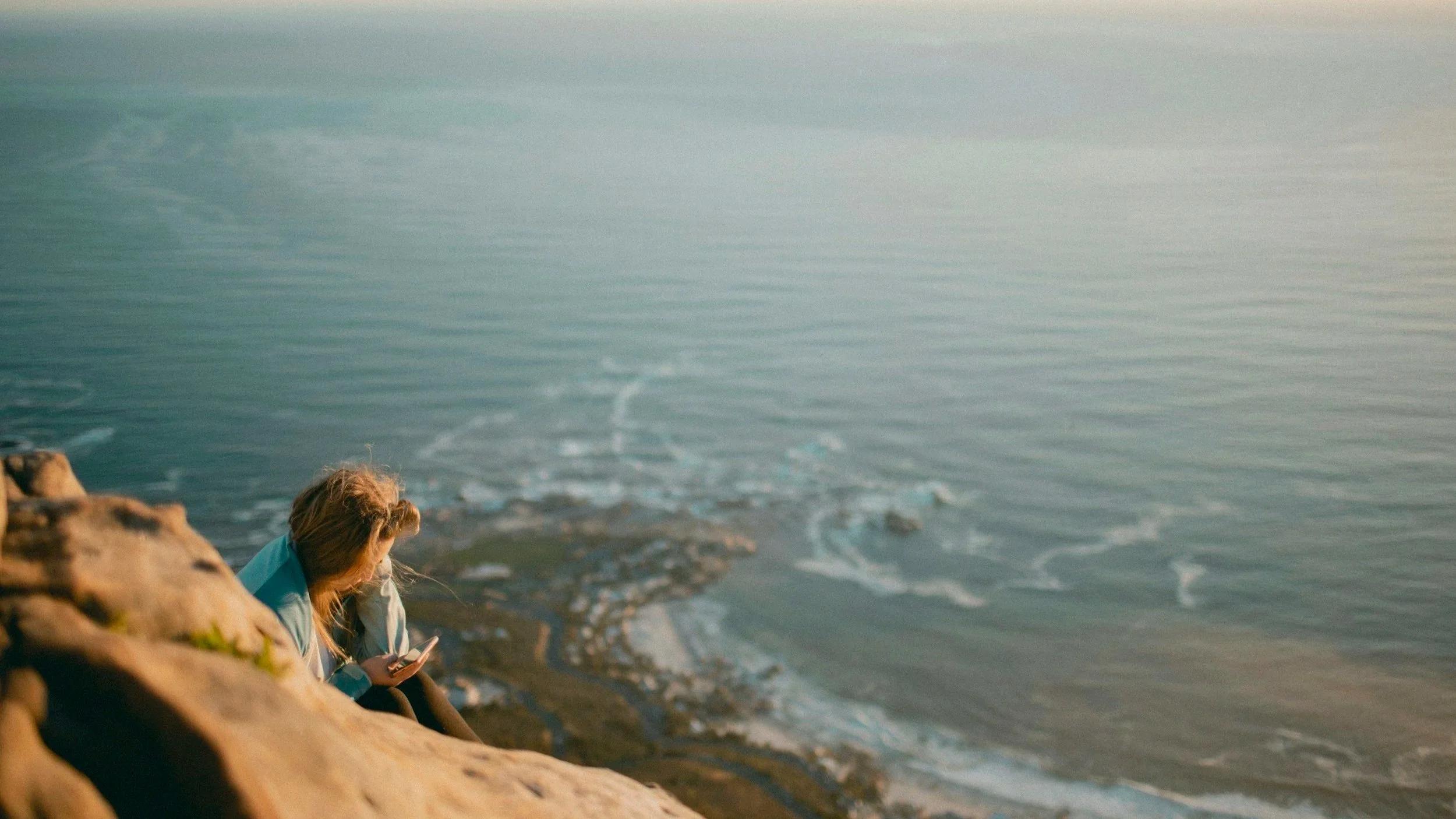 Mujer sentada sobre una roca, vestida con un pantalón y chamarra a una distancia elevada al lado de un paisaje con el mar viendo su teléfono celular.
