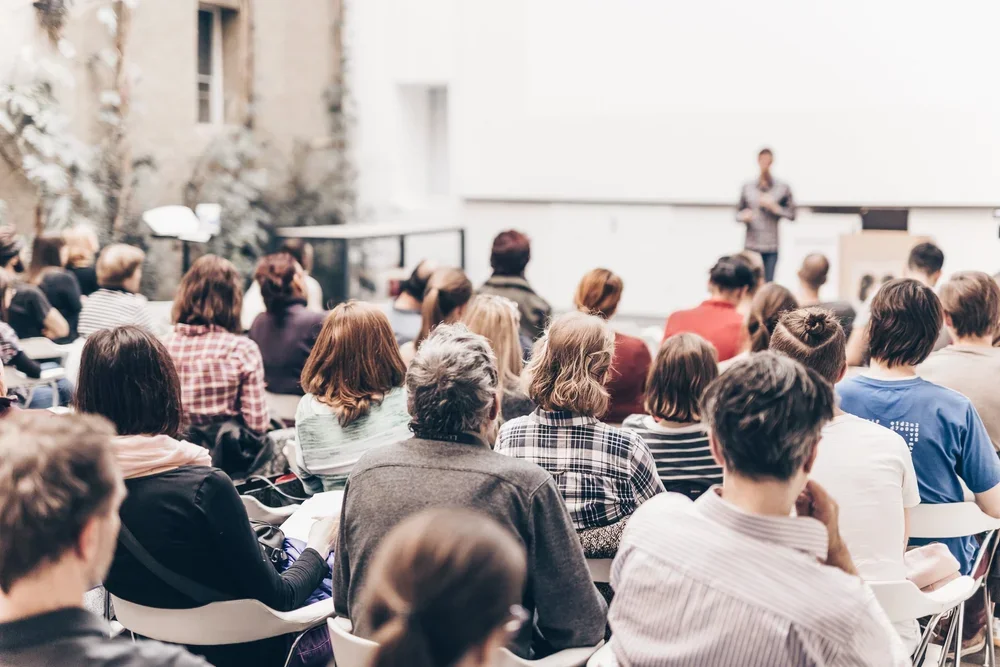 Auditorio lleno de personas asistiendo a una conferencia con un comunicador de pie frente a una pizarra compartiendo información crucial de Semiología de la Vida Cotidiana para transformar sus vidas.