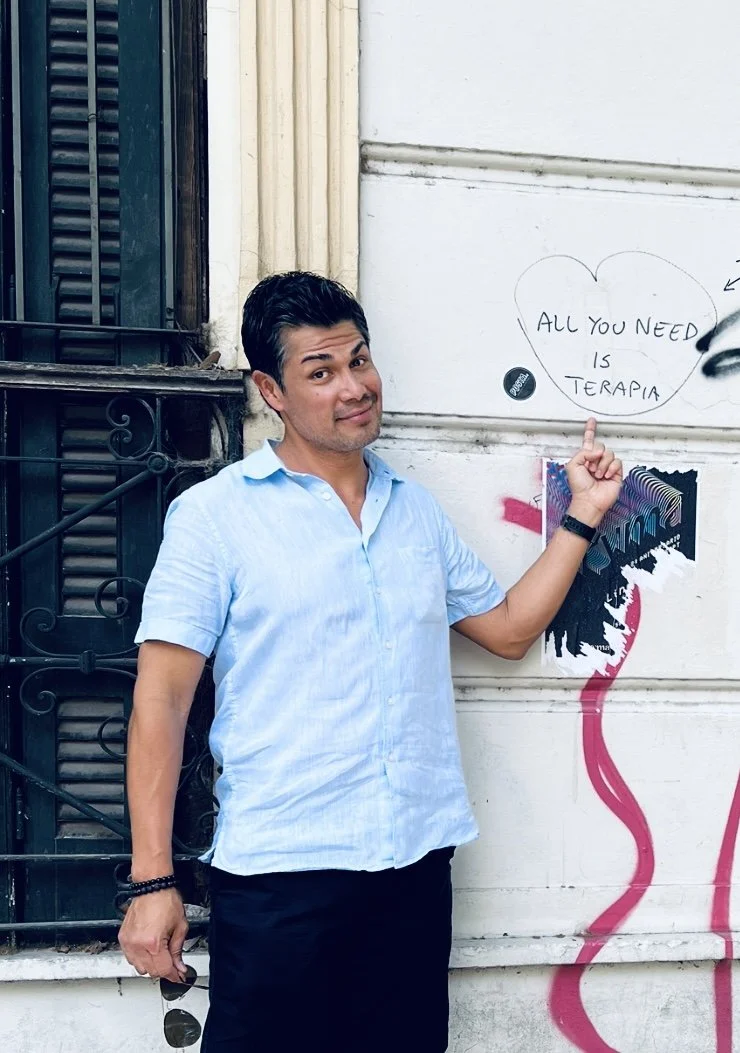 Foto de Arturo Sánchez Conócete a ti mismo.com con camisa azul claro apunta a una pared con graffiti y un mensaje que dice 'Todo lo que necesitas es terapia' en un globo de diálogo con forma de corazón. No es terapia en español. Es educador de vidas.