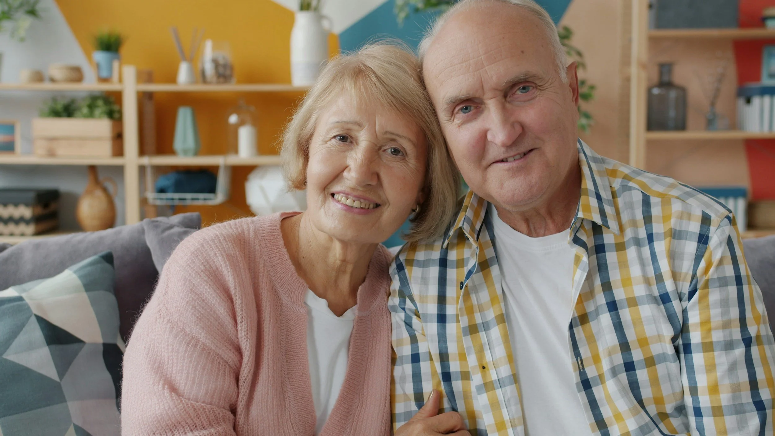 Older couple smiling, sitting close together on a gray couch with patterned pillows in a colorful, modern living room.