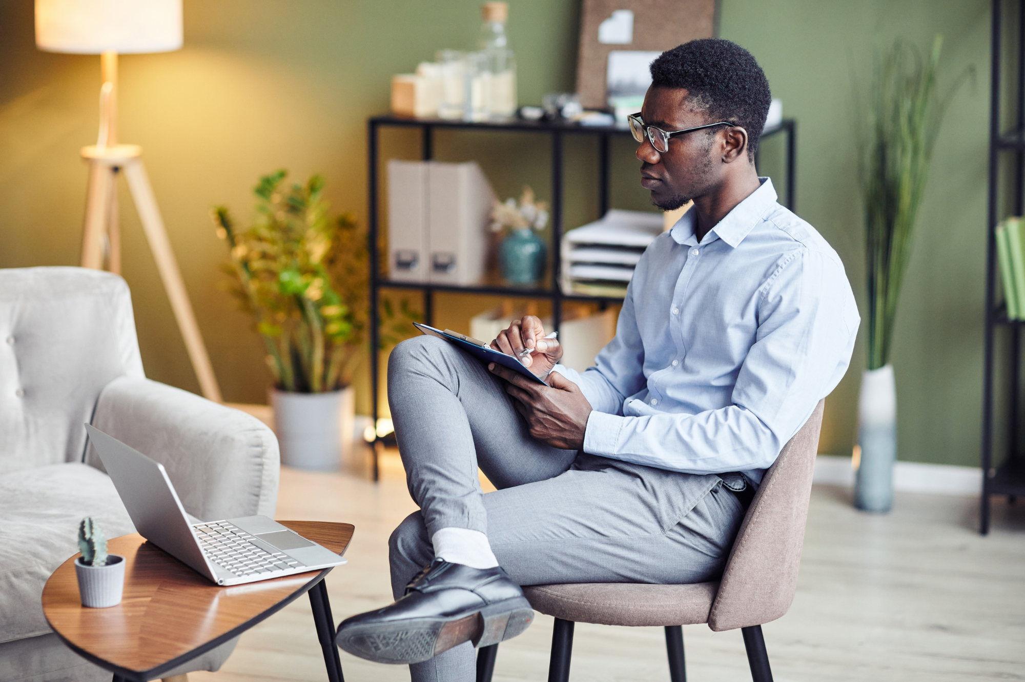 A man sitting on a chair in a modern office, taking notes on a tablet, with a laptop and a small cactus plant on a wooden table nearby, and green walls with shelves and plants in the background.