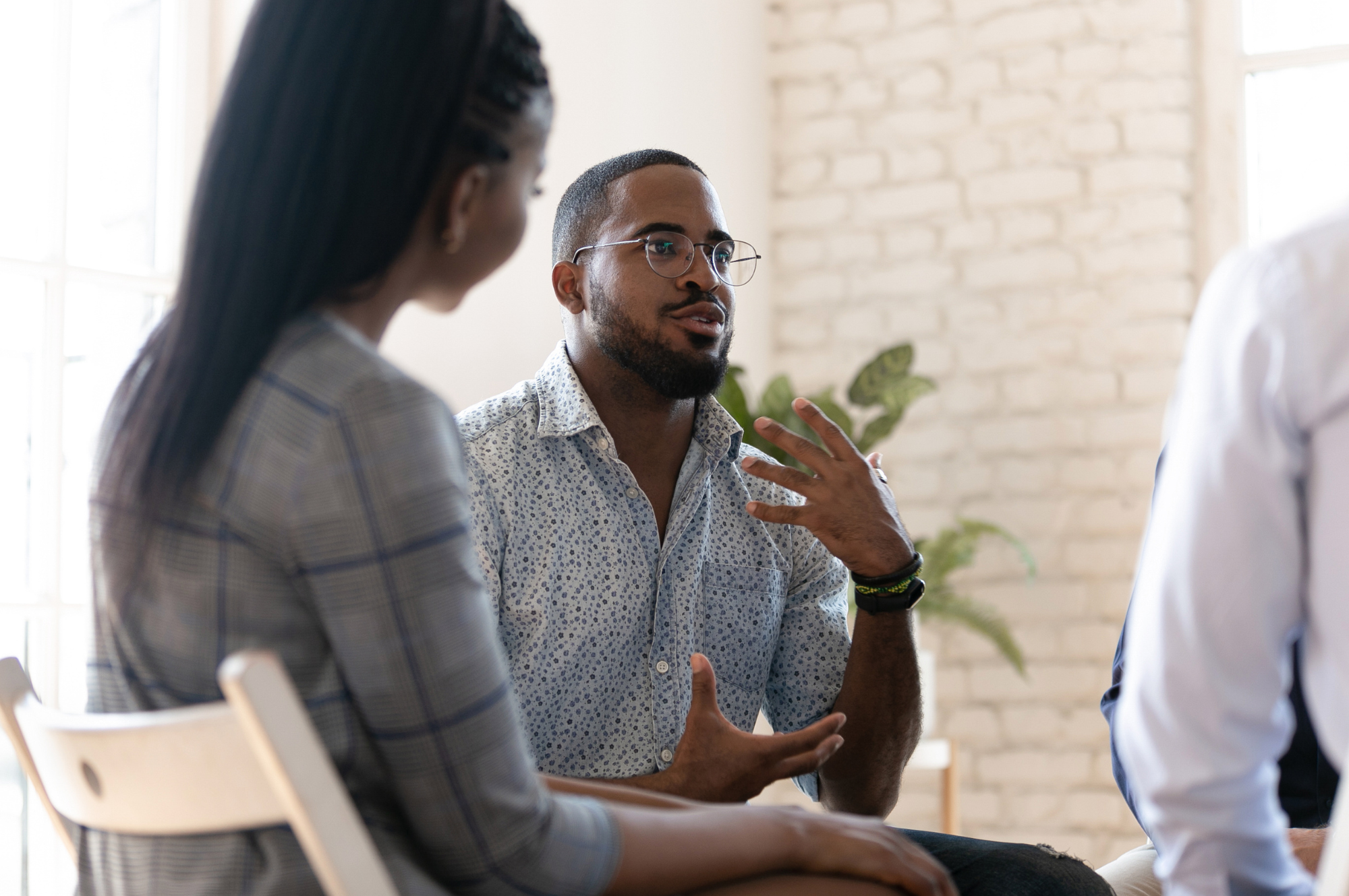 Three people engaged in a conversation indoors, with a brick wall and a window in the background. The man in the center is speaking, gesturing with his hands, and wearing glasses and a patterned shirt.
