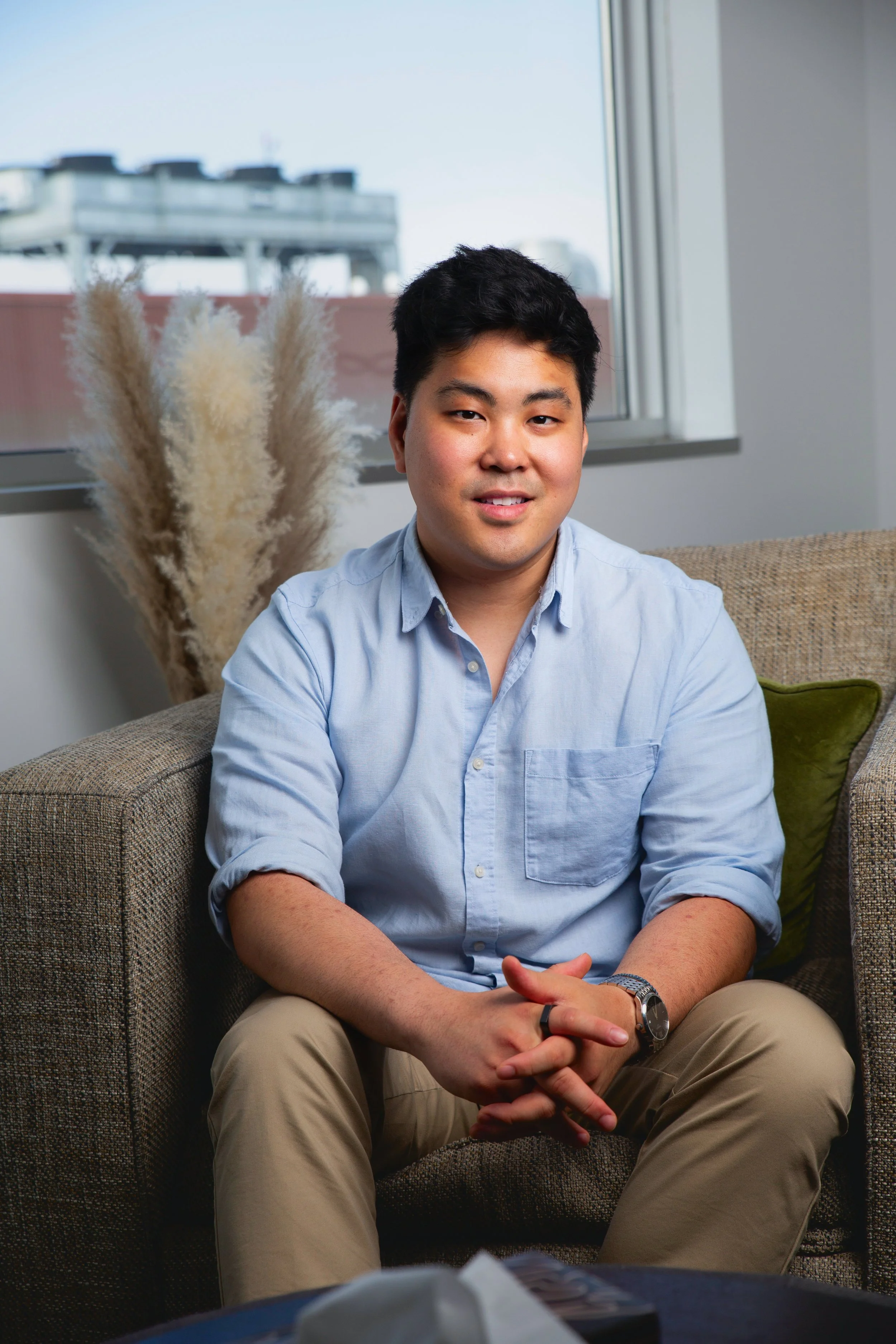 A young man with black hair sitting on a brown couch in a room with a window, beige curtains, and a green pillow, smiling at the camera. There are dried pampas grass in the background.