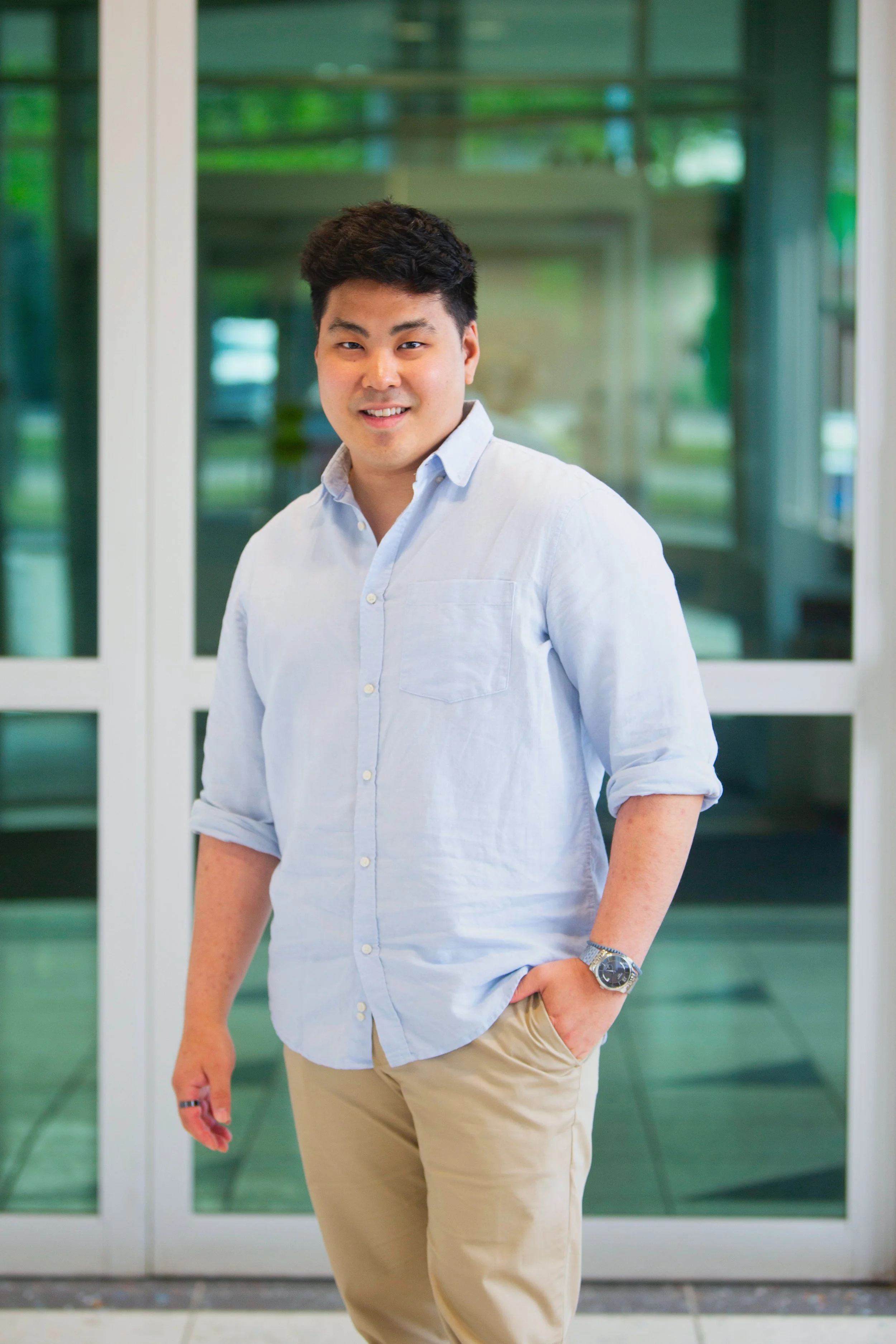 A young man with dark hair and wearing a light blue button-up shirt and khaki pants posed in front of glass doors.