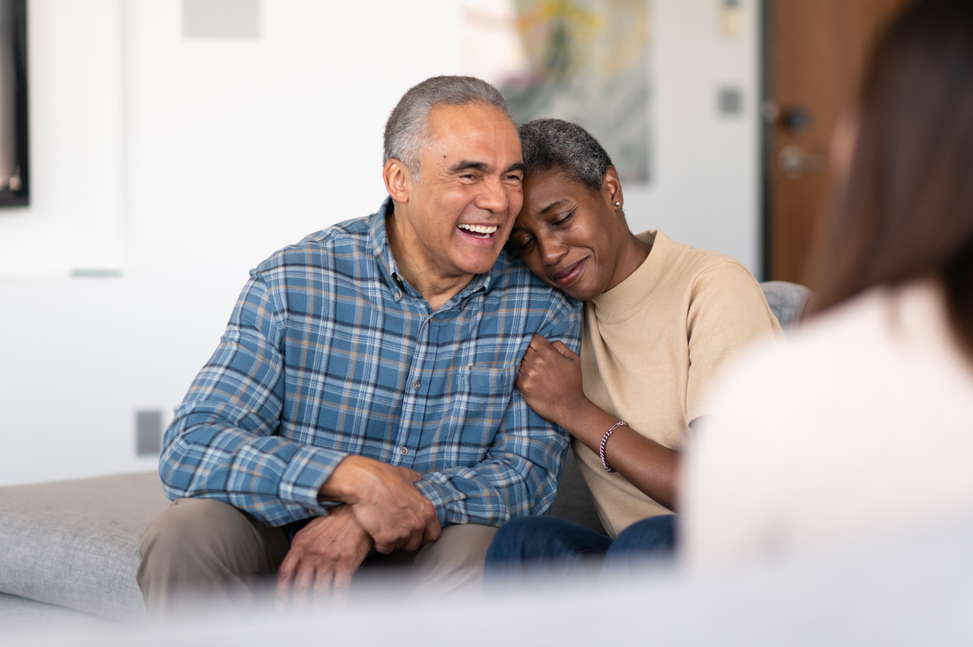 An elderly man and woman sharing a heartfelt moment during therapy or counseling session, sitting closely together on a sofa with a therapist in the foreground.