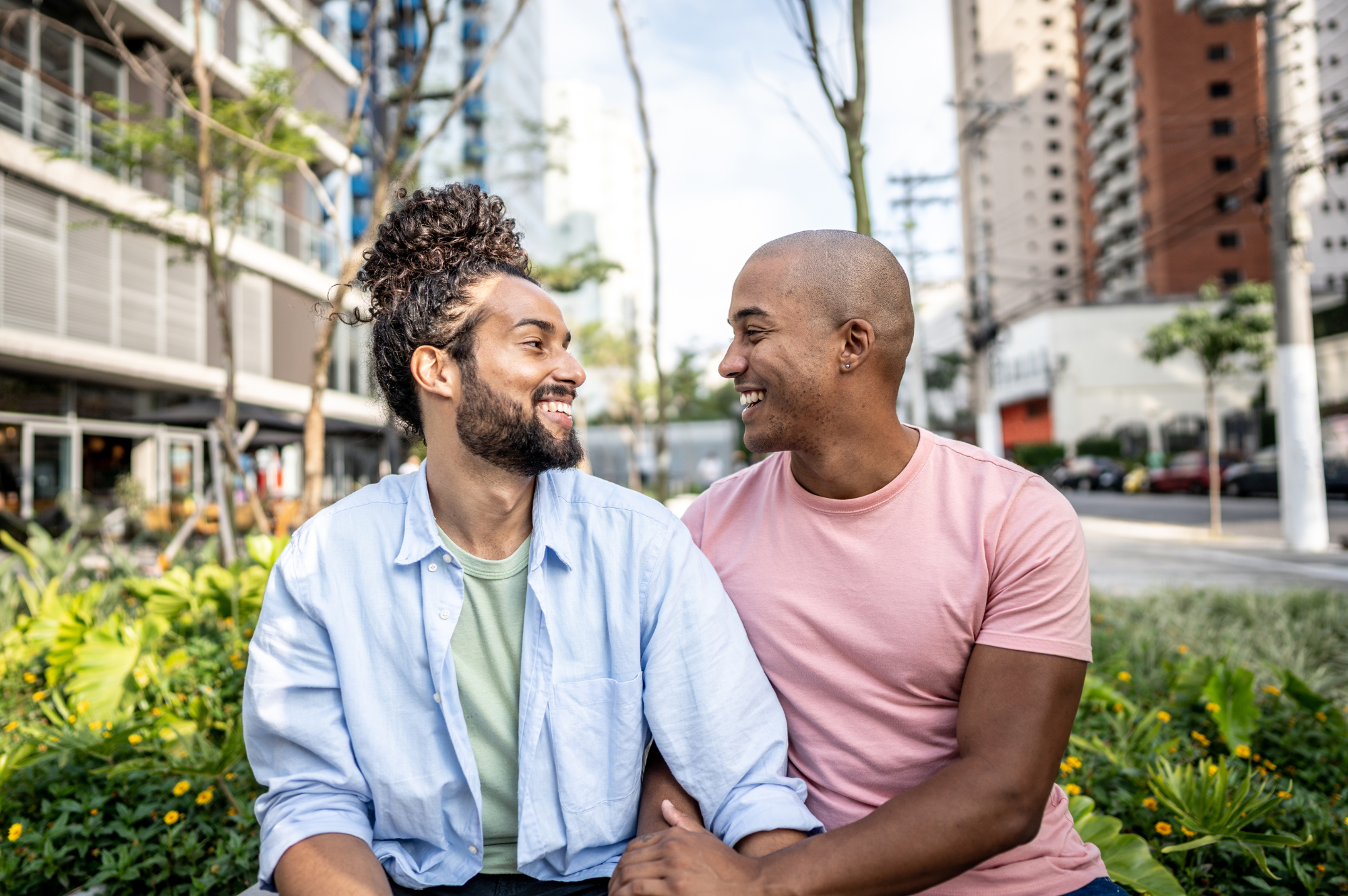 Two men sitting close together outdoors, smiling and looking at each other, in an urban park with buildings and trees in the background.